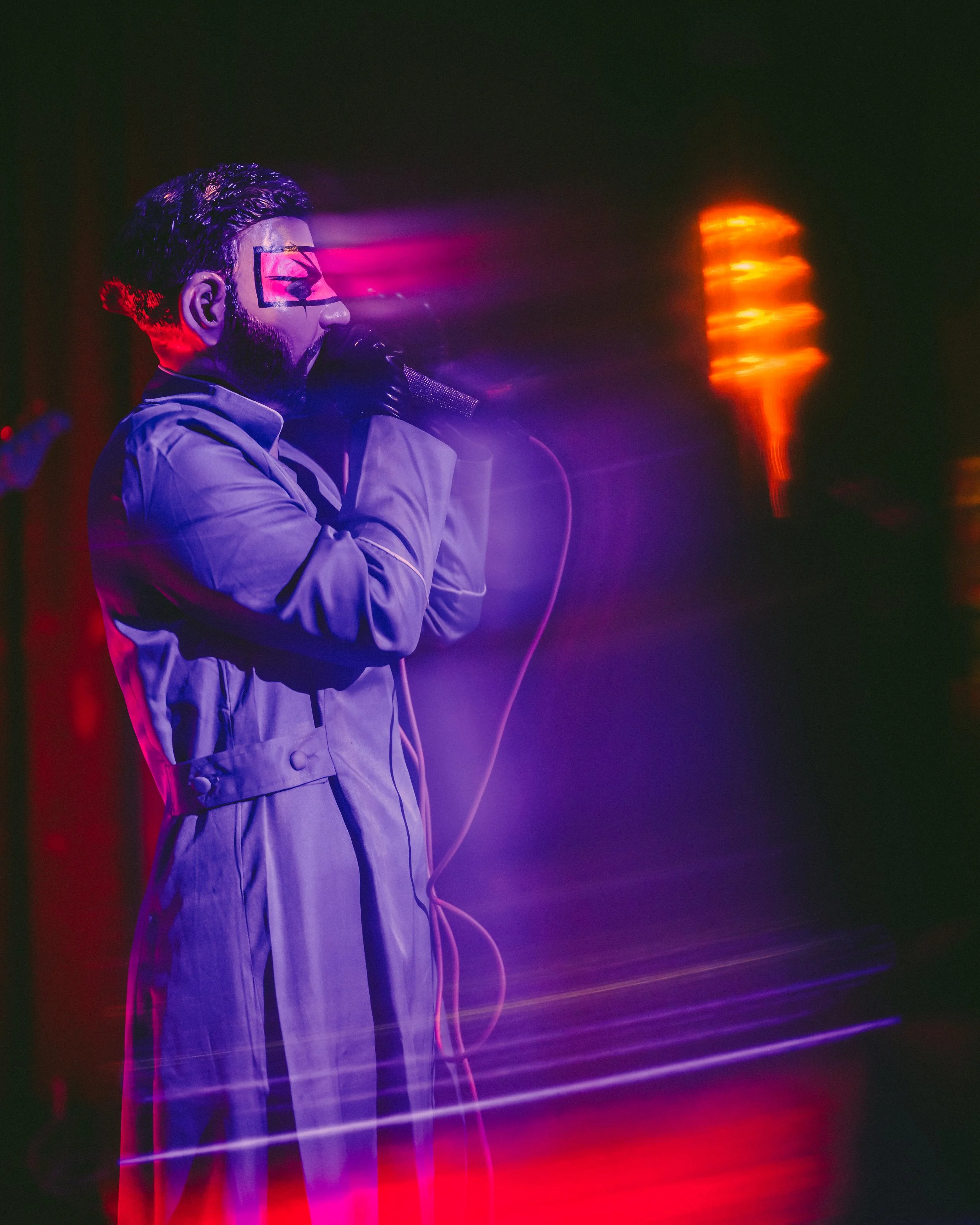 A person with face paint, wearing a gray jacket and gloves, holding a microphone on stage with colorful light effects.