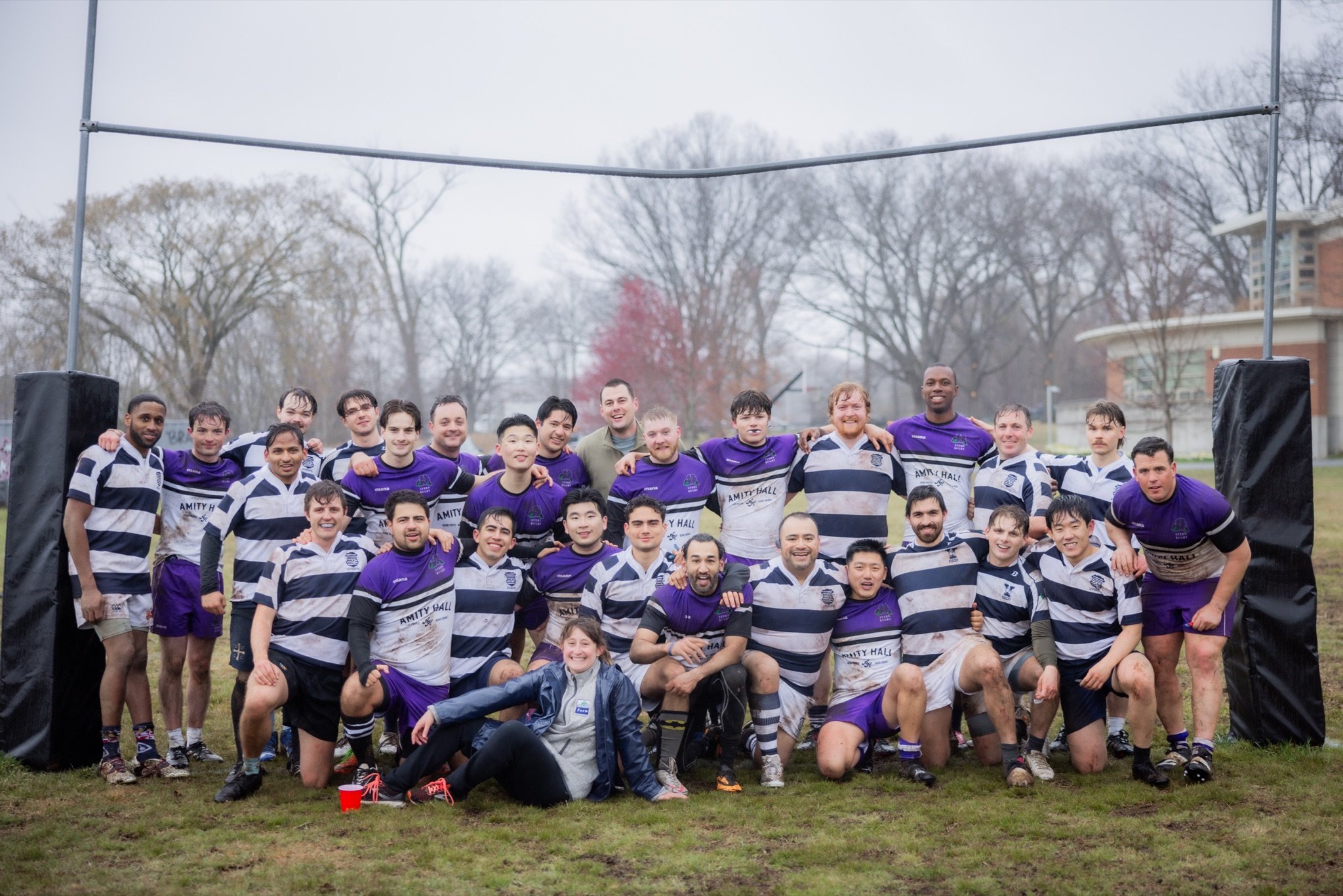 A rugby team group photo with about 23 players and 2 coaches, outdoors on a muddy field, wearing purple and black uniforms, after a game, with a goalpost behind them and leafless trees in the background.