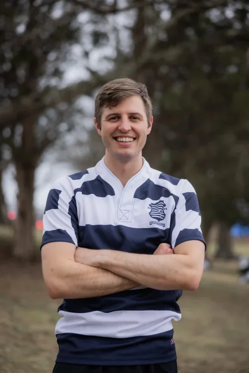 A smiling young man stands outdoors with arms crossed, wearing a navy and white striped sports jersey with a crest on the left side, amidst trees.