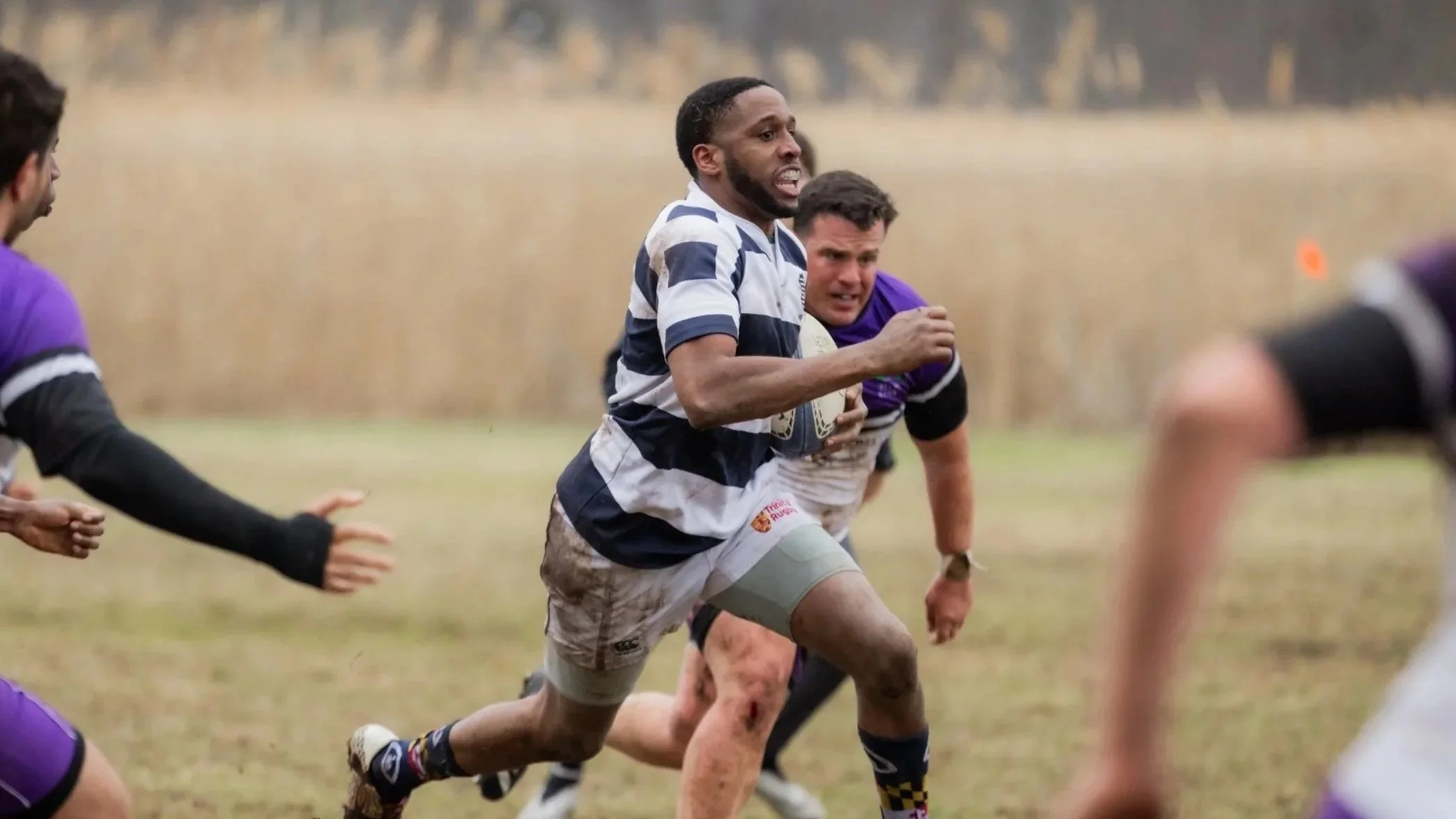 Rugby players in action during a match, with one player sprinting forward holding a rugby ball while others pursue him, on a grassy field with blurry background.