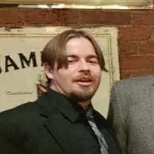 A man with light brown hair and a beard wearing a black suit and a tie, standing in front of a brick wall with a sign that partially reads 'JAM'.