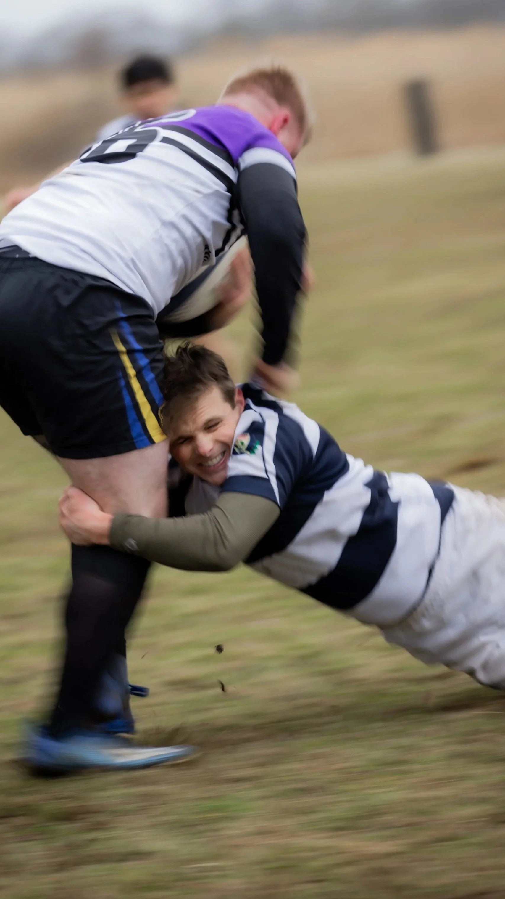 Two rugby players in a tackle on a grassy field, with one player smiling as he is tackled to the ground.