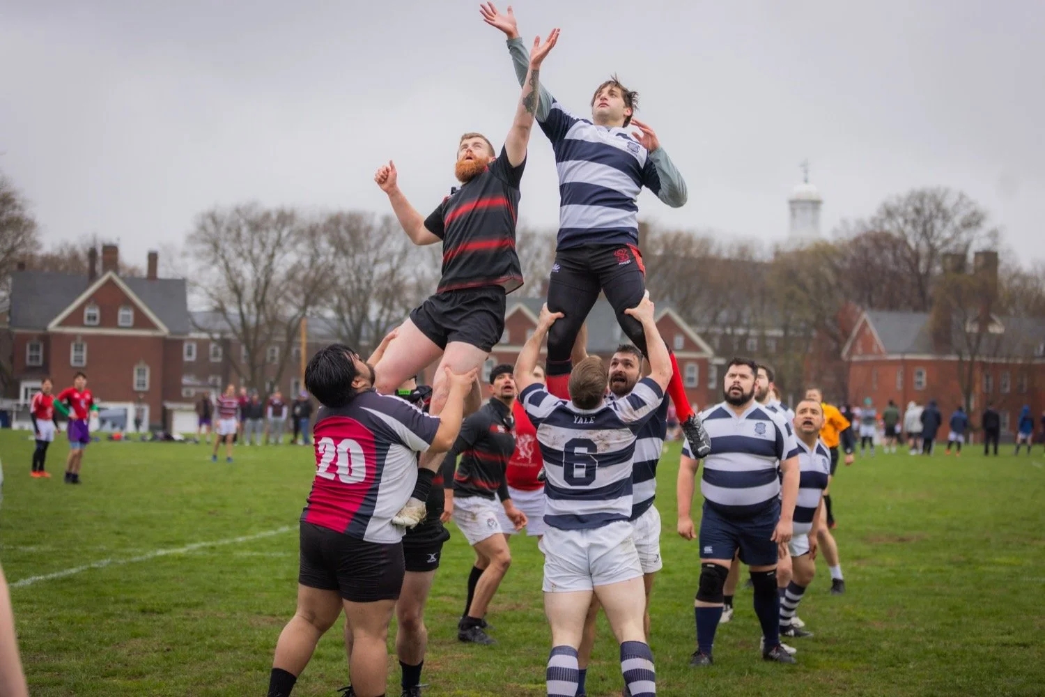 Rugby players in a lineout contest, lifting one player to catch the ball, on a grassy field with spectators and houses in the background.