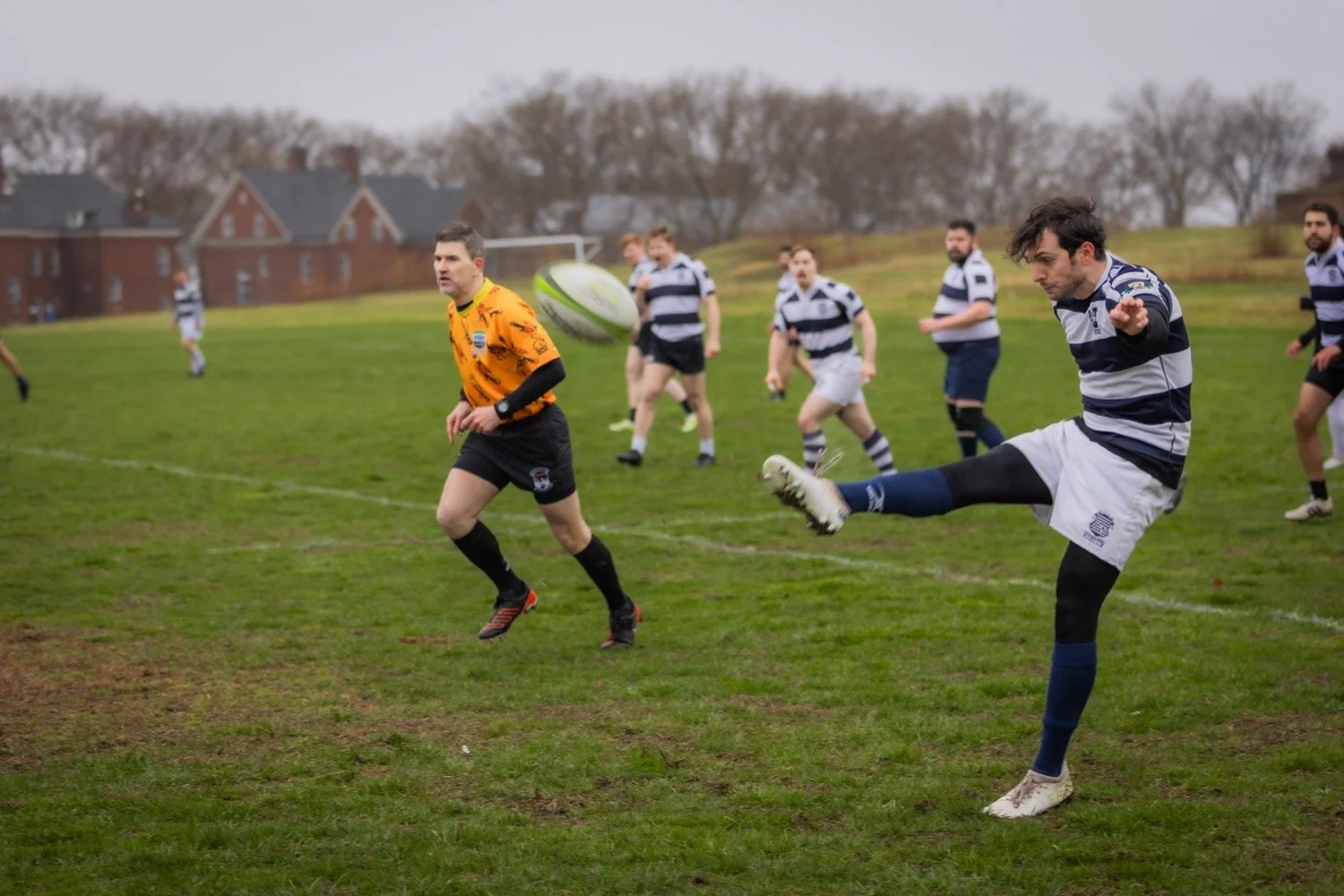 A rugby match with players on a grassy field. One player in a blue and white striped uniform kicks the ball, while others in the background, including a referee in a yellow shirt, watch and play.