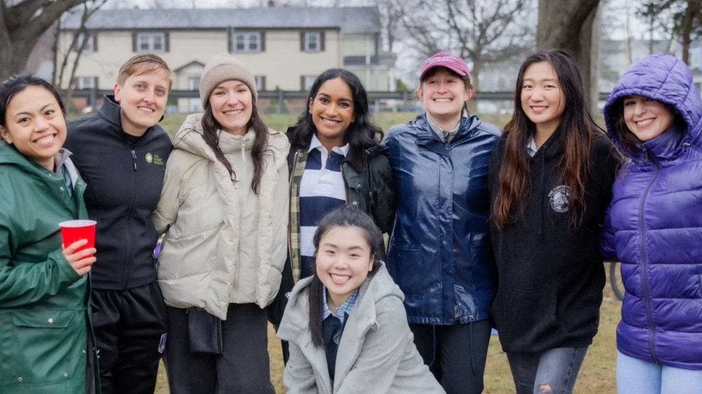 Group of eight diverse young women smiling outdoors on a cloudy day, wearing jackets and hoodies, standing close together.