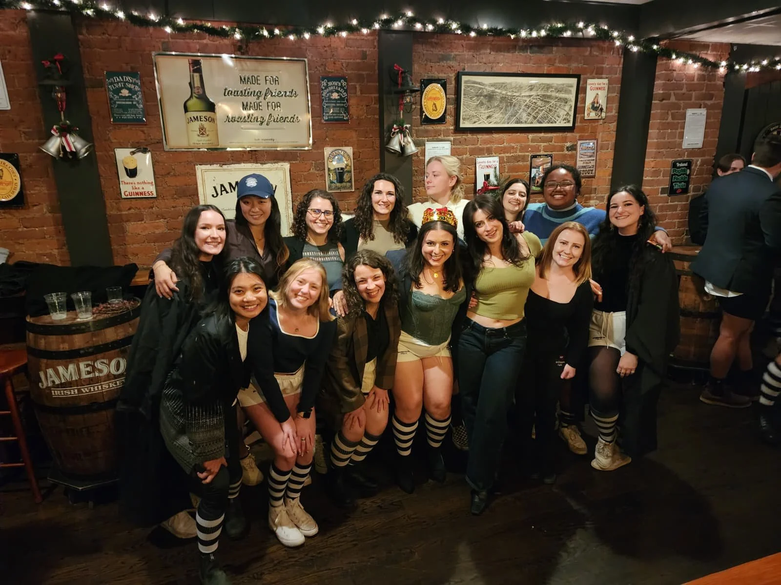 A group of women celebrating together at a bar, some wearing festive headbands and striped socks, with brick walls and framed posters in the background.