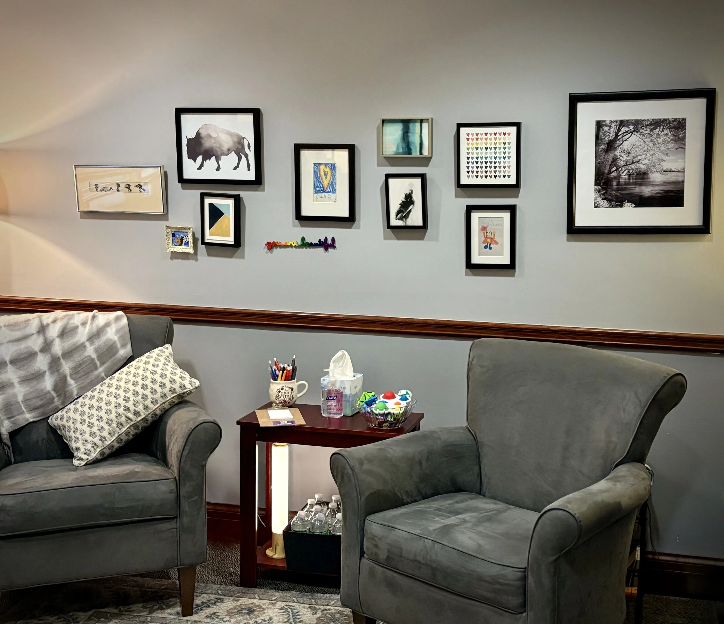 A cozy living room with two gray armchairs, a small wooden side table, and a gallery wall of framed artwork on the wall. The paintings include black and white photography, abstract art, and colorful illustrations.