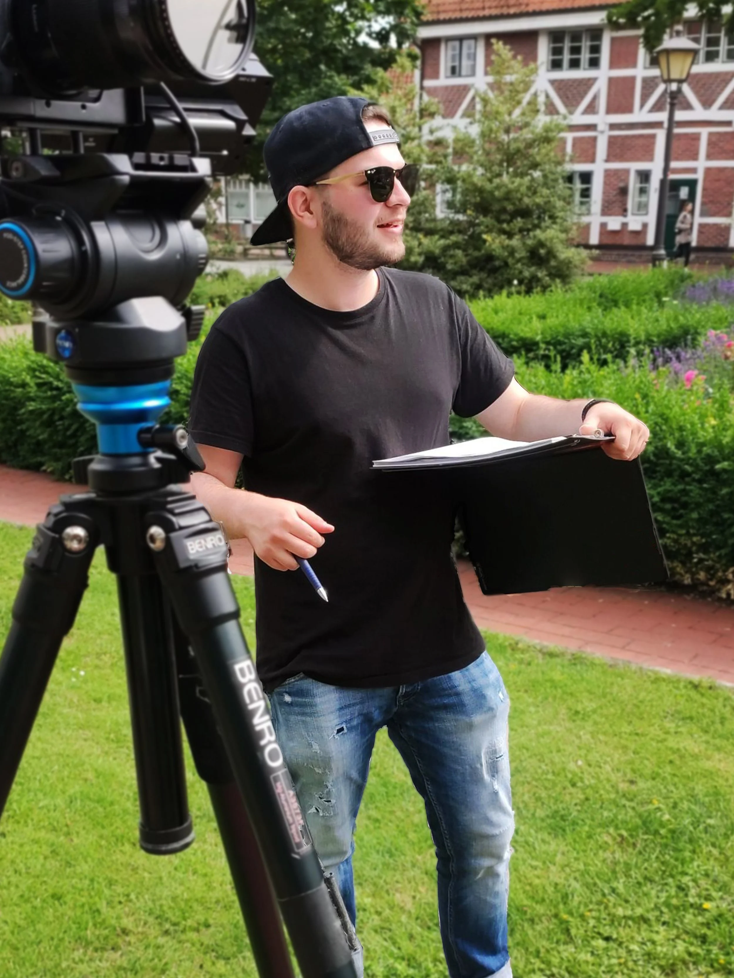 Ein Mann mit Sonnenbrille und Basecap steht neben einer Kamera auf einem Stativ in einem grünen Garten (Vor dem Rathaus der Gemeinde Jork). Er hält Klemmbrett und Stift in der Hand. Im Hintergrund ist ein Fachwerkhaus sichtbar. (Moritz Fürste)