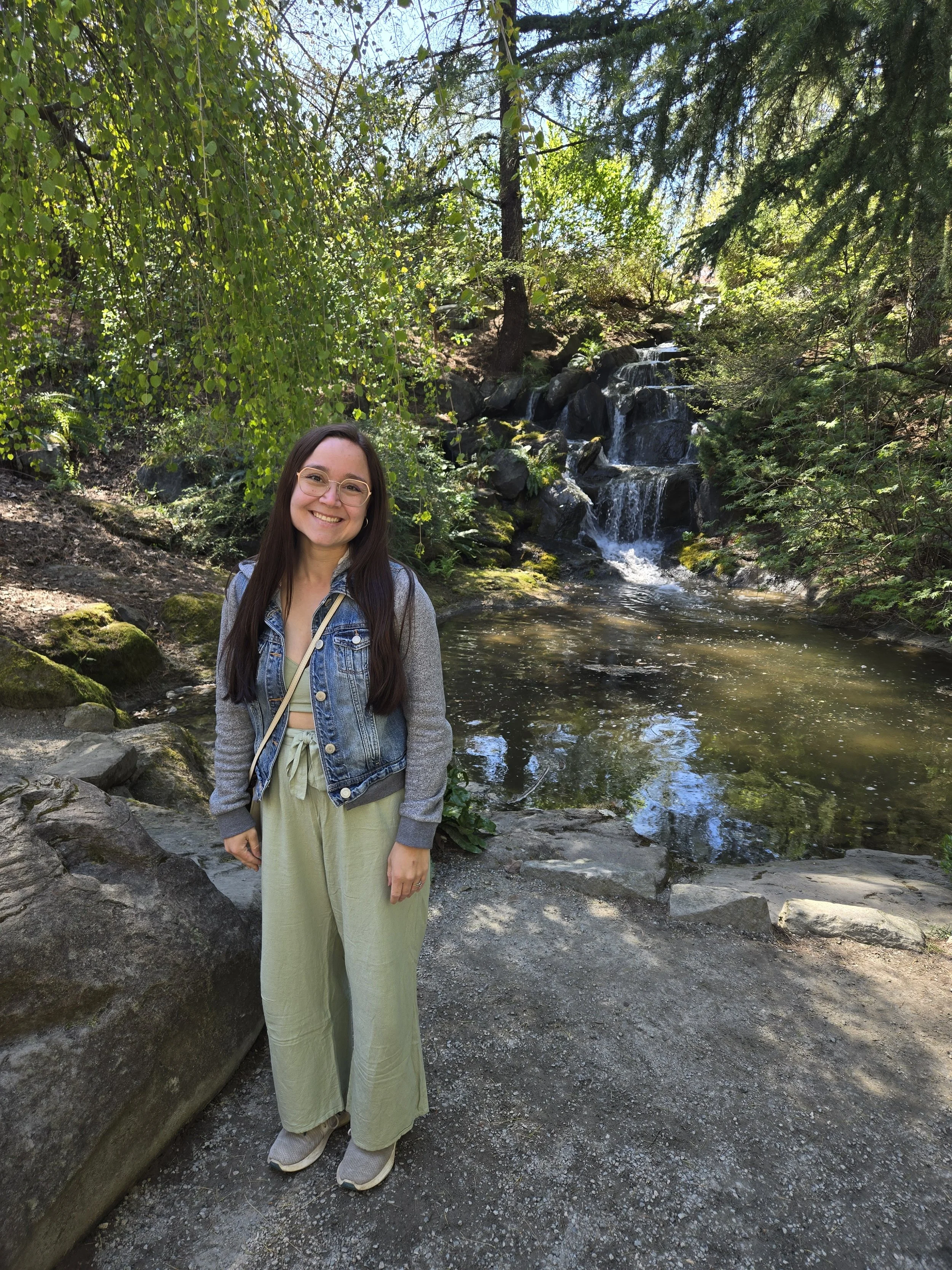 A smiling woman with long dark hair, glasses, wearing a denim jacket, sage pants, and sneakers standing in front of a small waterfall and pond in a lush, green forest.