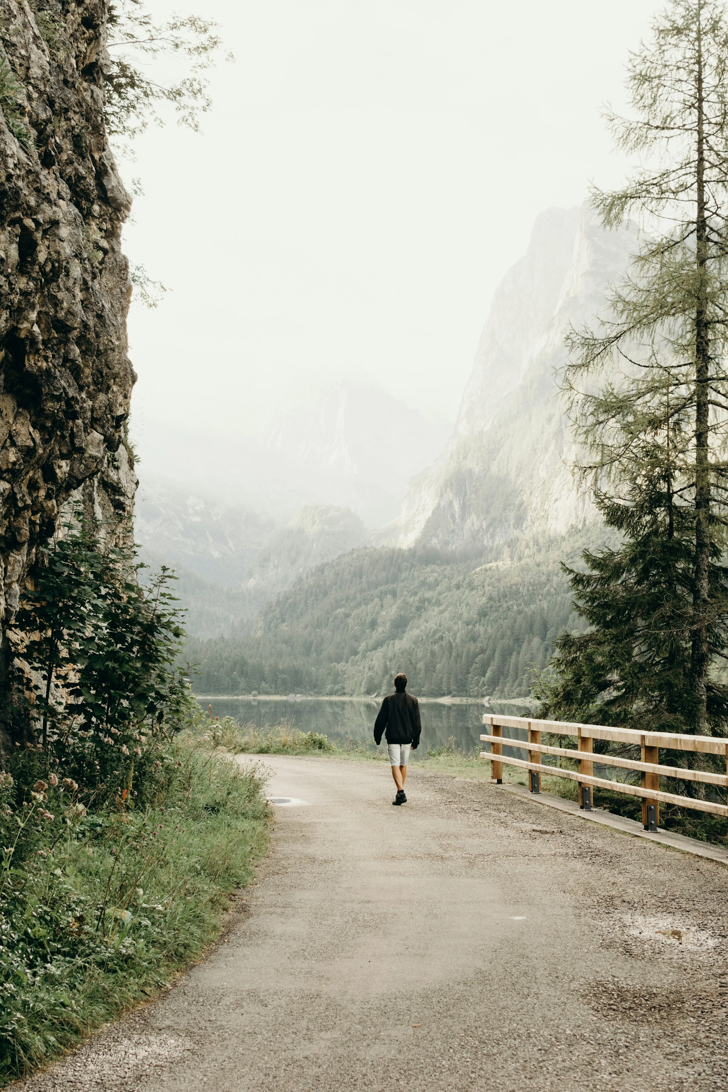 A person walking on a gravel path beside a lake, surrounded by mountains and trees, with overcast weather.