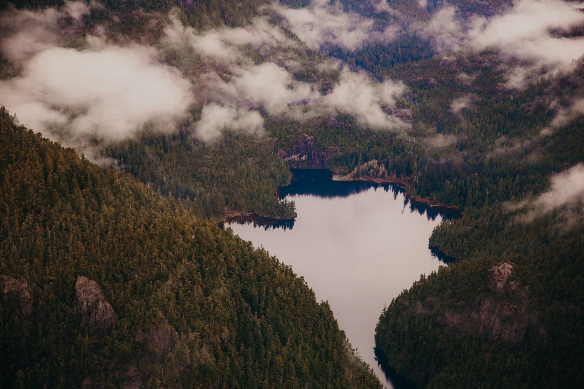 Aerial view of a large lake surrounded by dense forested mountains with clouds and fog overhead.