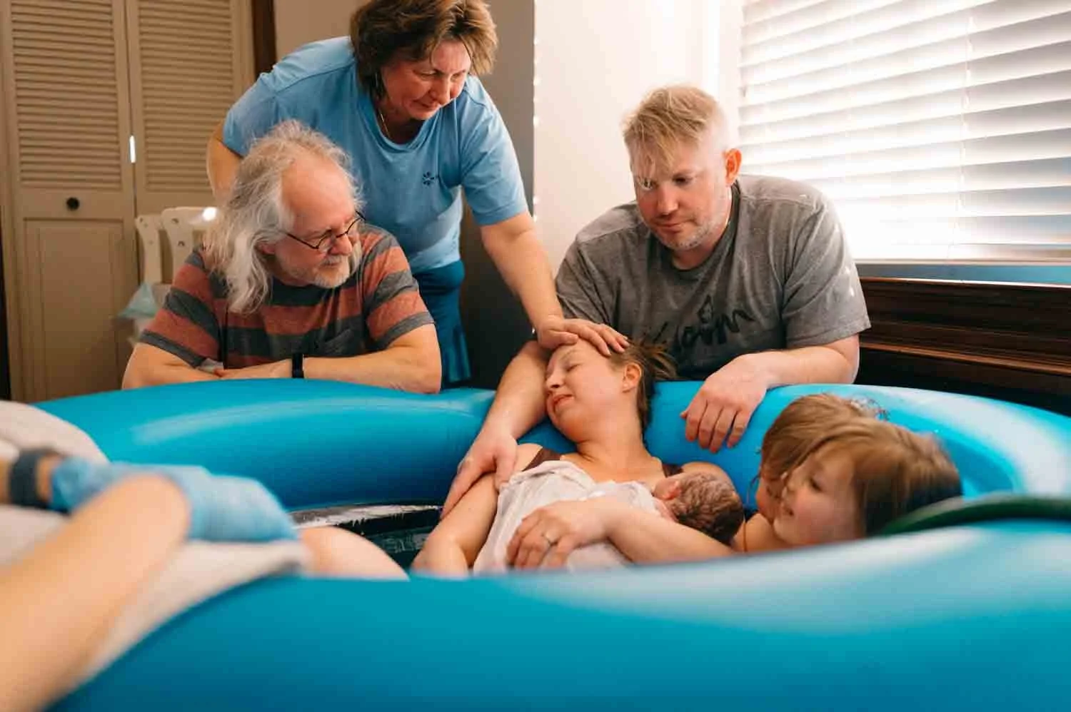 A family gathered around a woman in a birthing pool, who is holding a newborn baby. They appear relaxed and joyous, with natural lighting through a window.