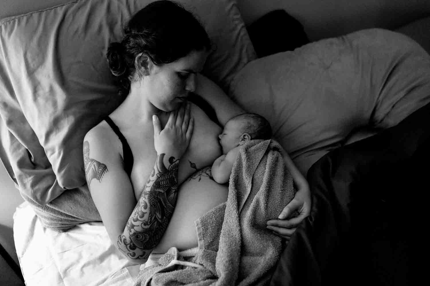 A black and white photo of a mother with tattoos holding her newborn baby while lying on a bed, covered with a towel.