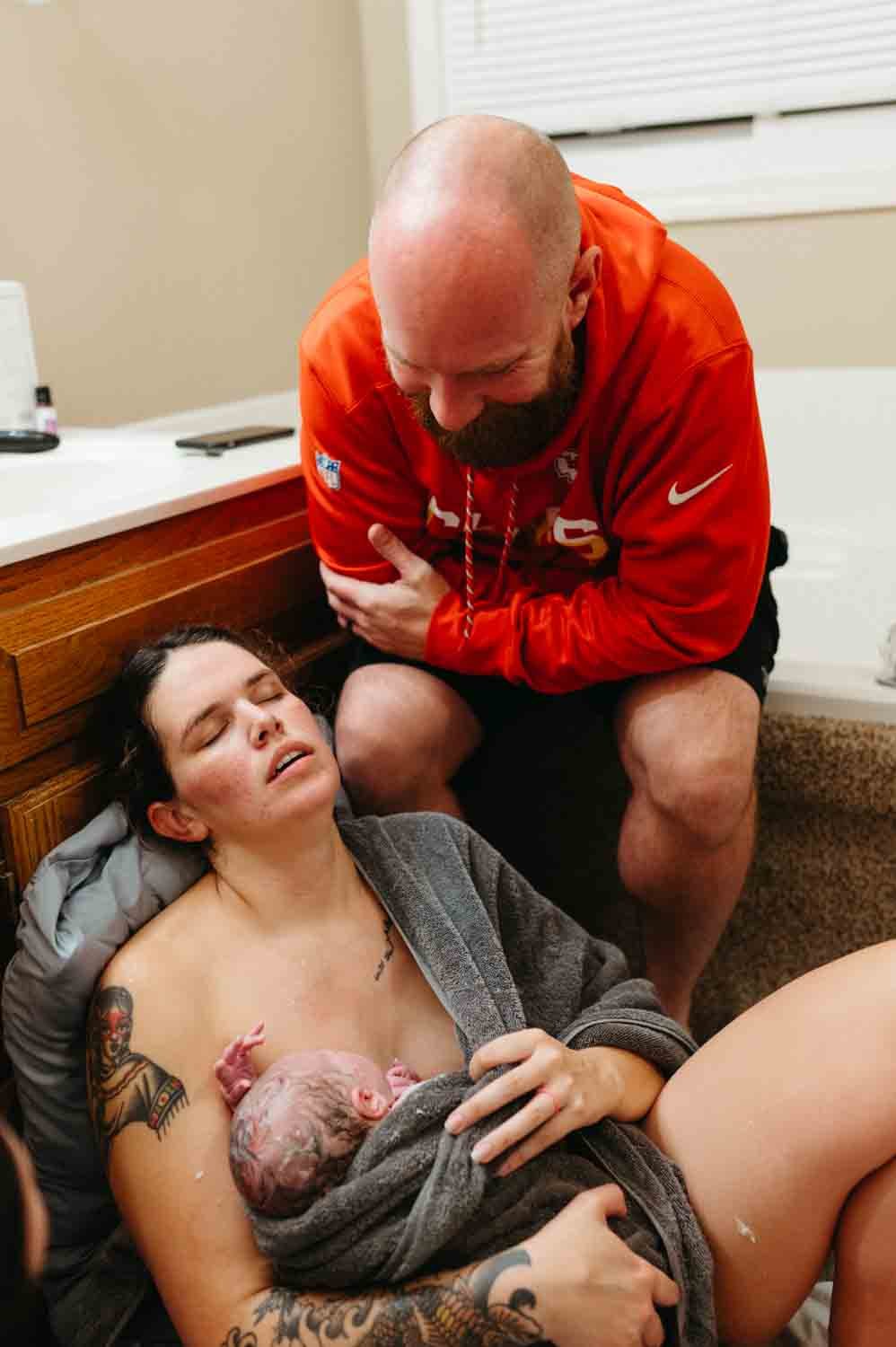 A mother holding a newborn baby wrapped in a towel, with a supportive individual beside her, in a home setting after childbirth.