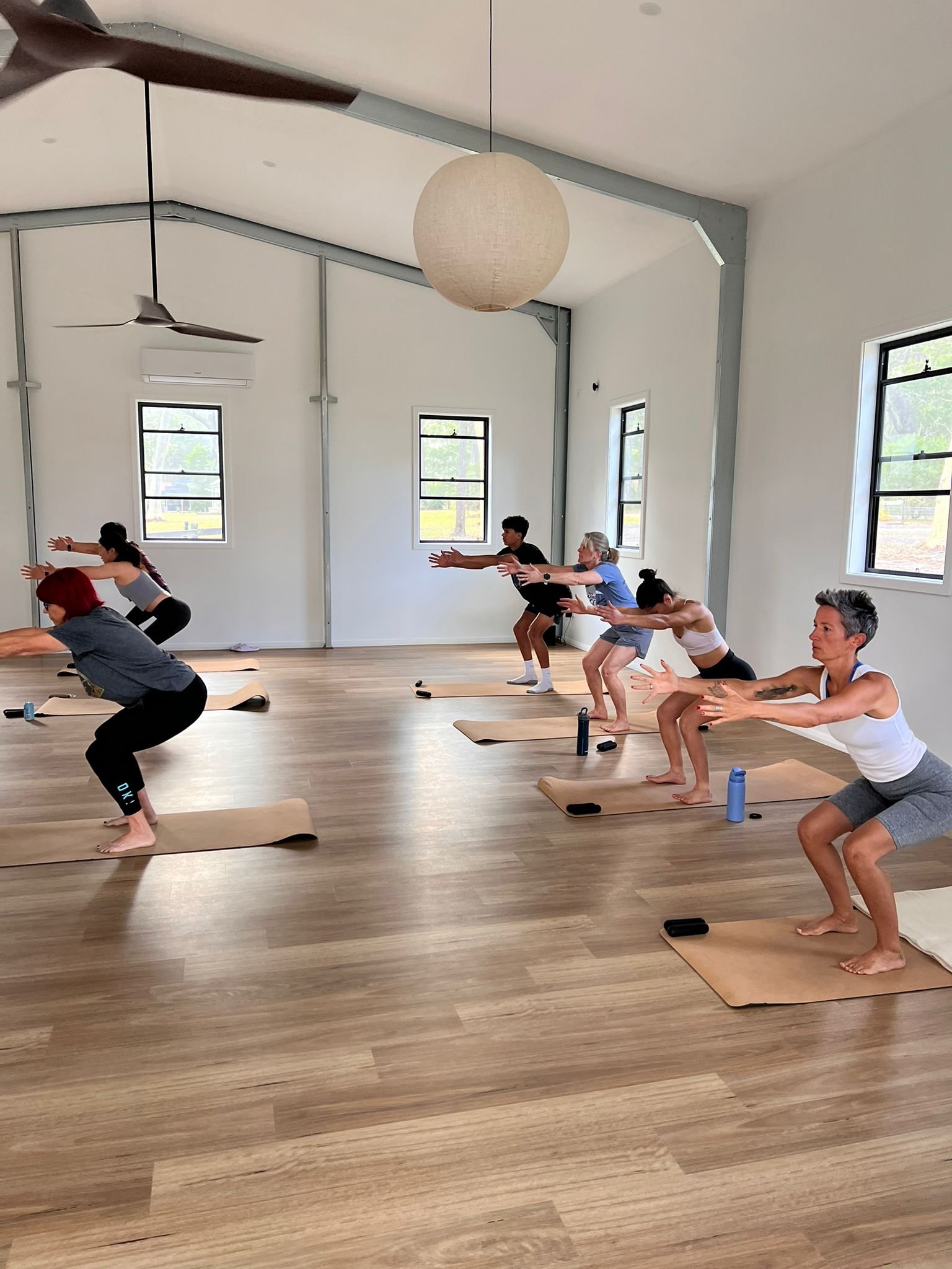 Group of people practicing yoga in a bright studio with wooden floors and windows, following an instructor.