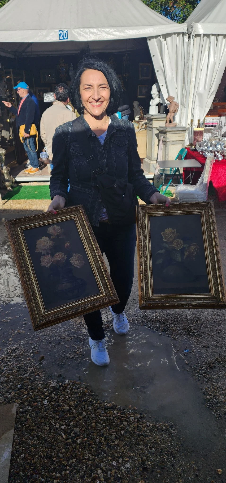 A smiling woman with black hair holding two framed floral paintings outdoors at a market. White tent and other people are visible in the background.
