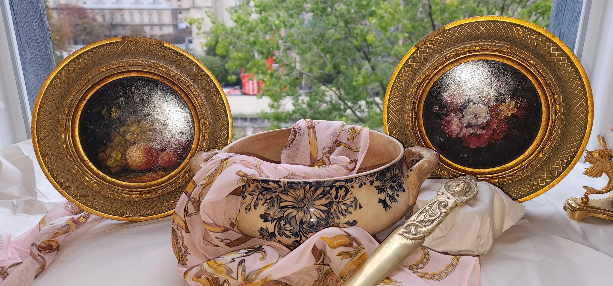 Decorative still life with two ornate gold framed paintings, a ceramic bowl with a floral design, pink patterned scarf, and a decorative gold object against a window with green trees outside.