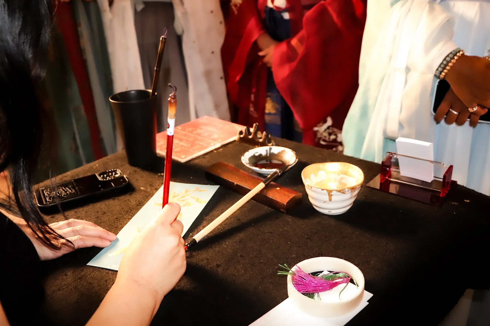 A person using a calligraphy brush at a table with ink and brushes, surrounded by other people, with various containers and art supplies on the table.