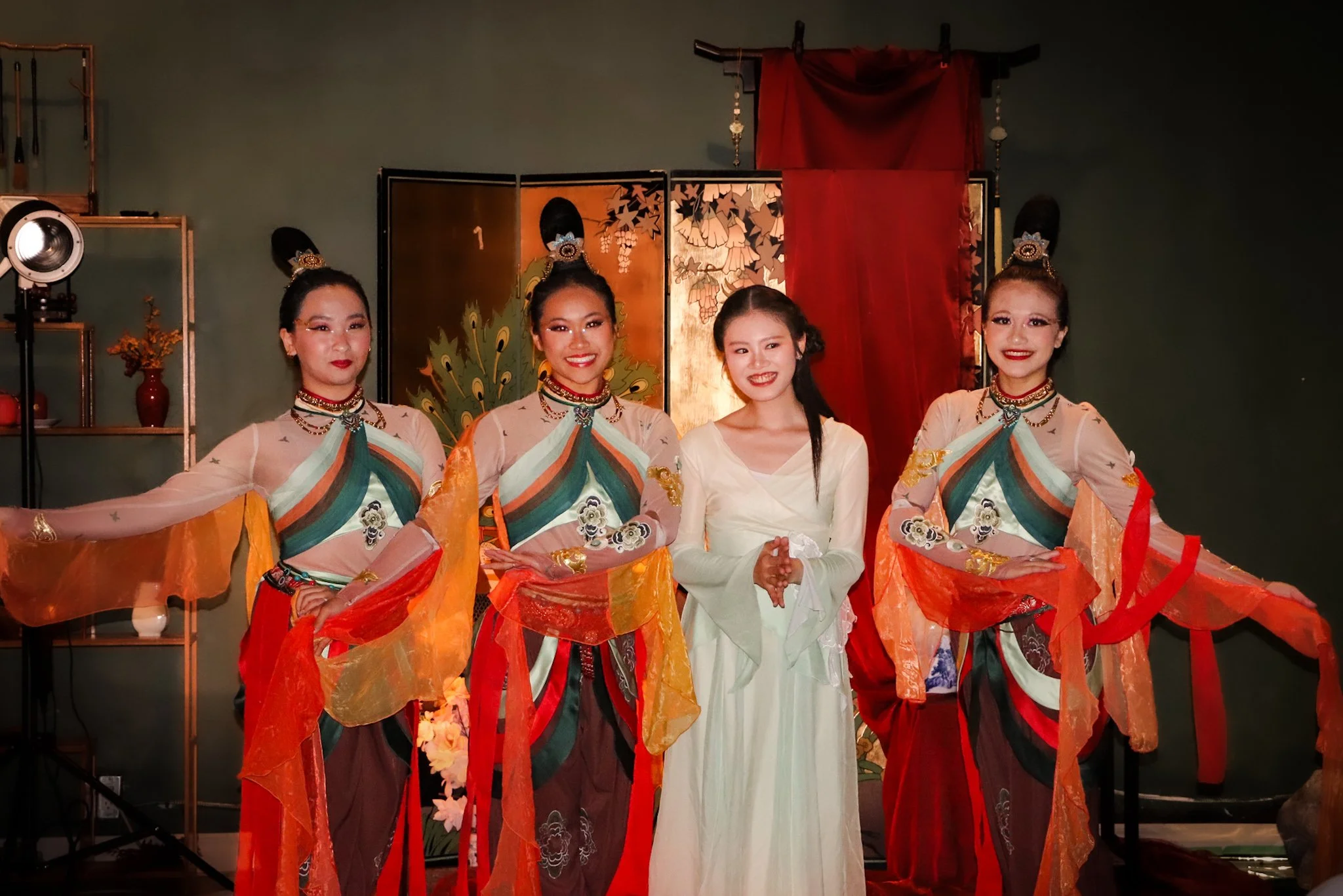 Four women dressed in traditional Asian costumes posing indoors, with a woman in the center wearing a white dress and three women on either side wearing colorful costumes with flowing sleeves and jewelry. Behind them is decorative art with red cloth 
