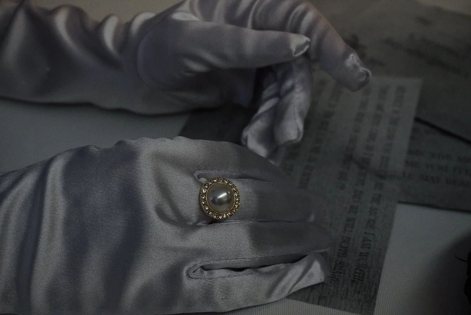 Close-up of a person's hand wearing gray gloves, showcasing a pearl ring with a gold border and embedded small diamonds.