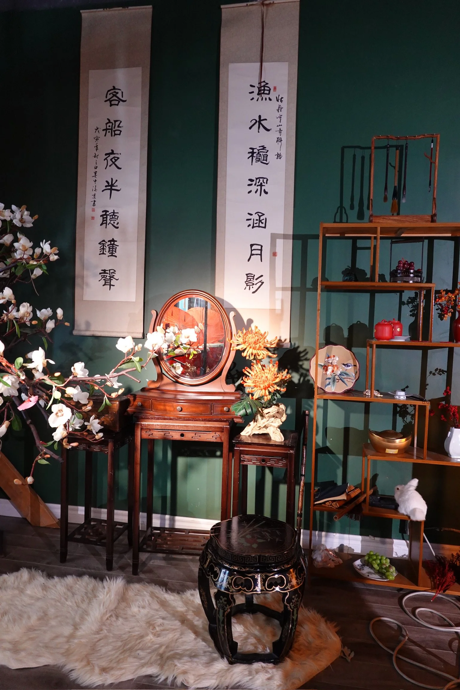An Asian-inspired interior decor with Chinese calligraphy scrolls on a green wall, a wooden vanity with a mirror, a vase with orange flowers, a black decorative stool, and various small ornamental objects on wooden shelves. There is a plush fur rug o