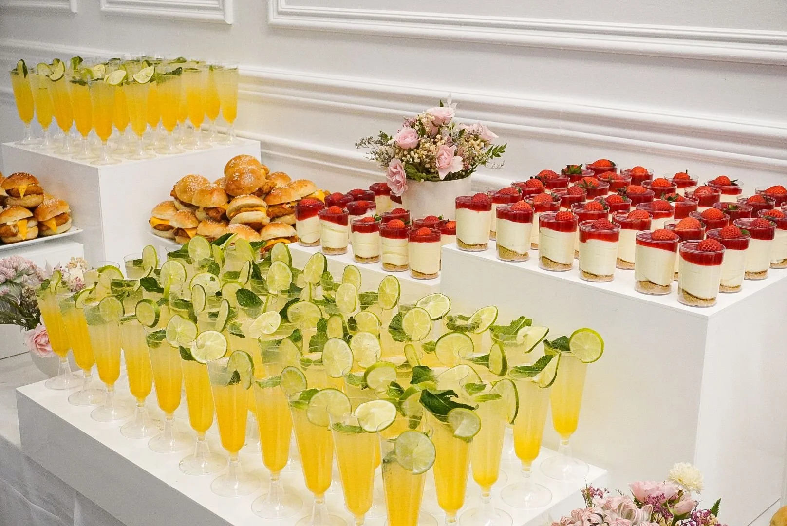 A dessert and drink table with lemon cocktails garnished with lime slices and mint, strawberry cheesecakes topped with strawberries, sliders with cheese and meat, and small glasses of creamy dessert topped with strawberries, decorated with pink flowe