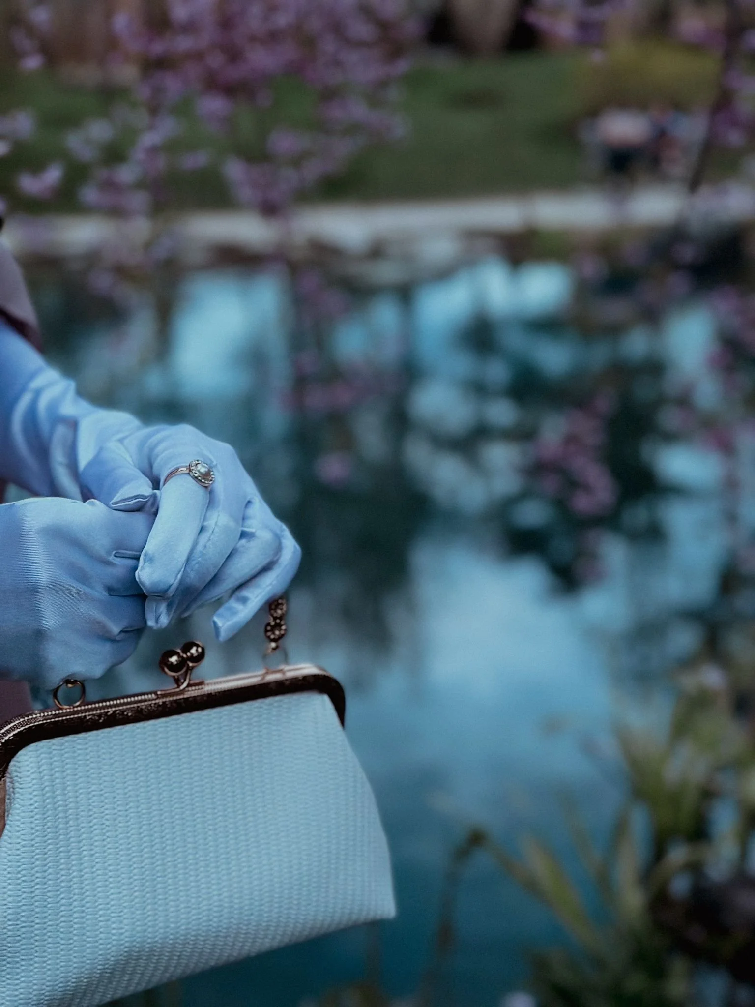 Close-up of a person wearing light blue gloves holding a small light blue purse with a metal clasp, with a blurred pond and pink flowering trees in the background.