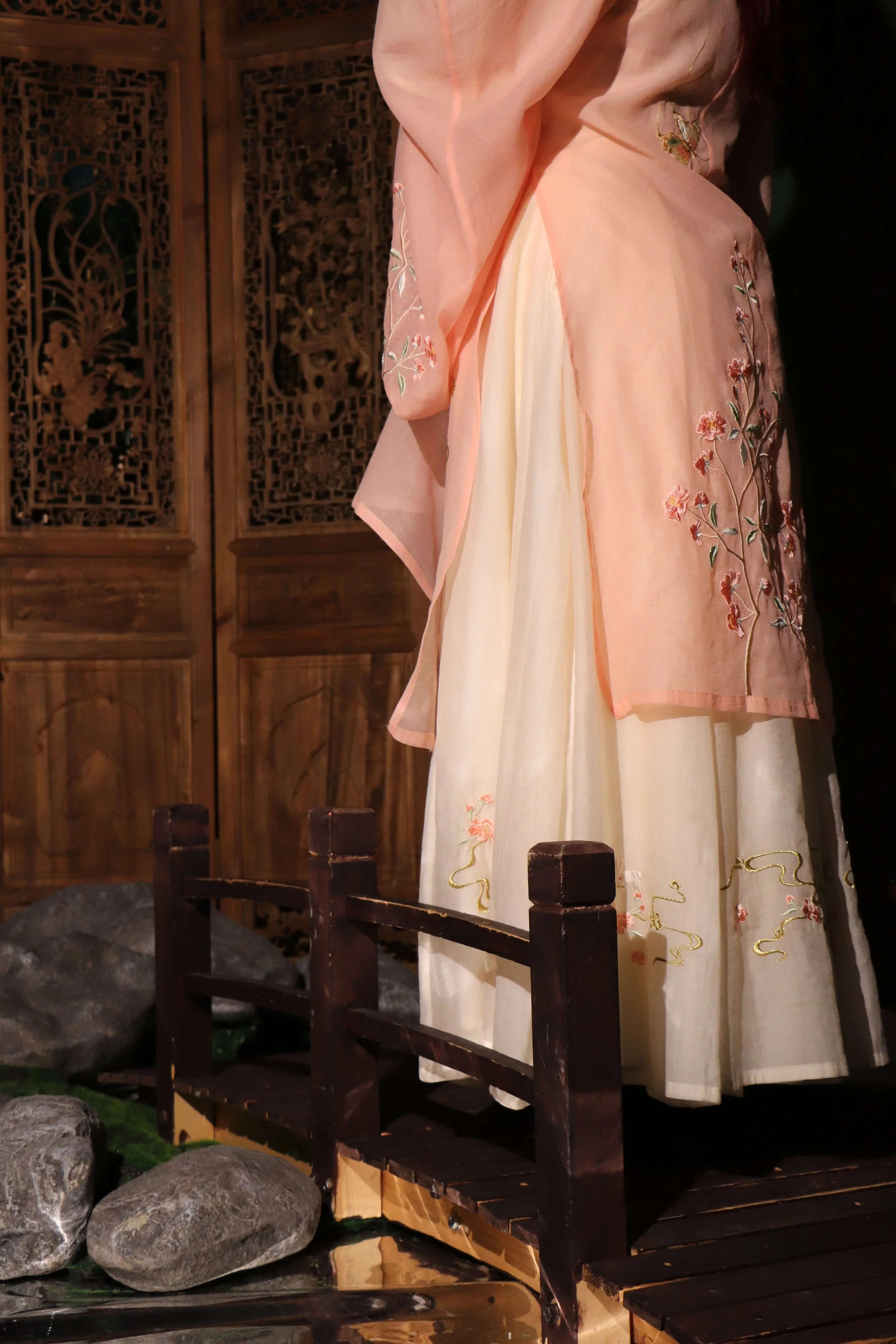 A traditional Korean hanbok with a pink jacket and a cream skirt with floral embroidery displayed on a mannequin. The background features wooden panels with intricate carvings, and the mannequin stands on a small wooden bridge over rocks.