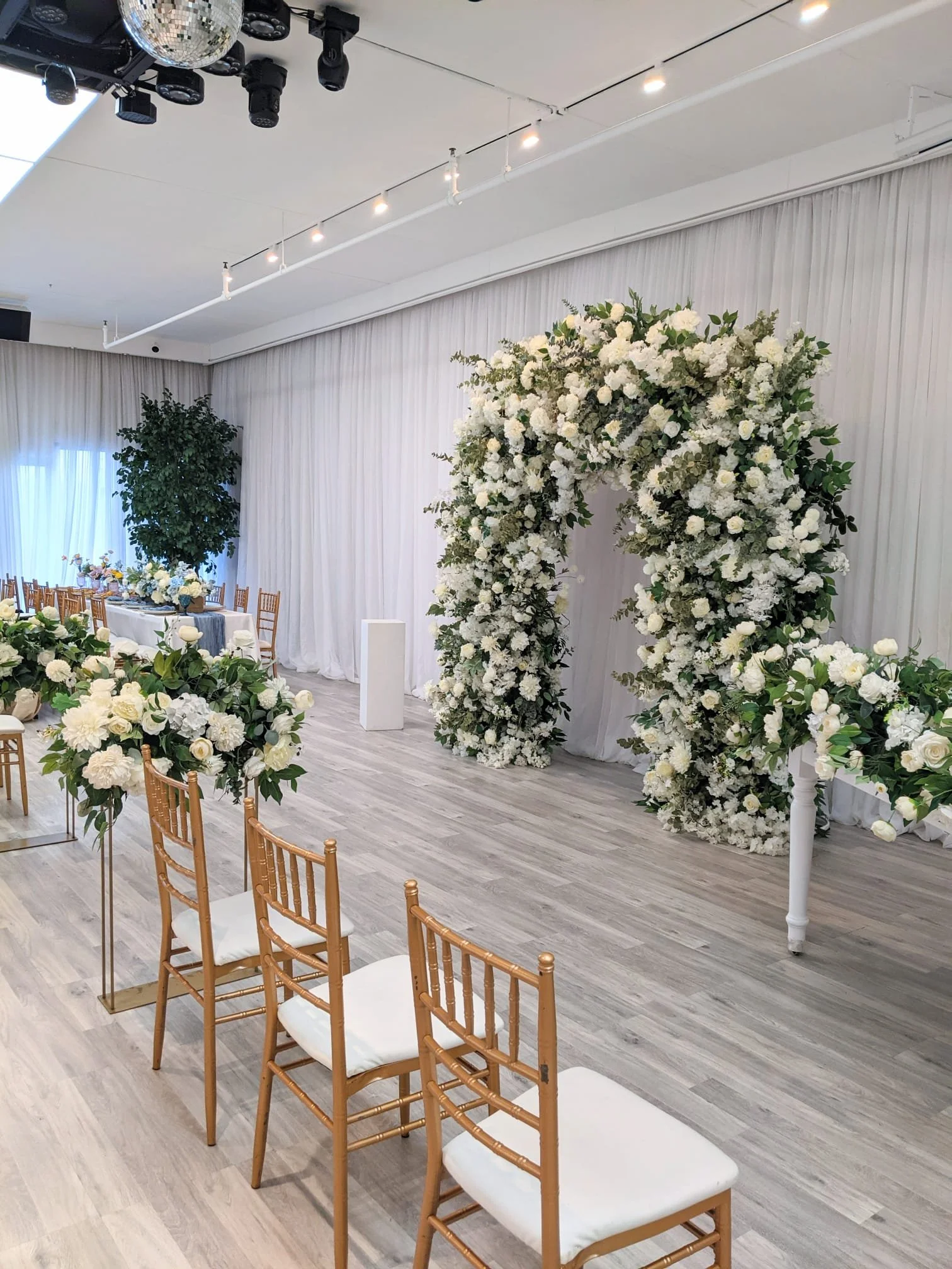 A decorated wedding venue with white floral arch, floral arrangements on tables, and rows of chairs.