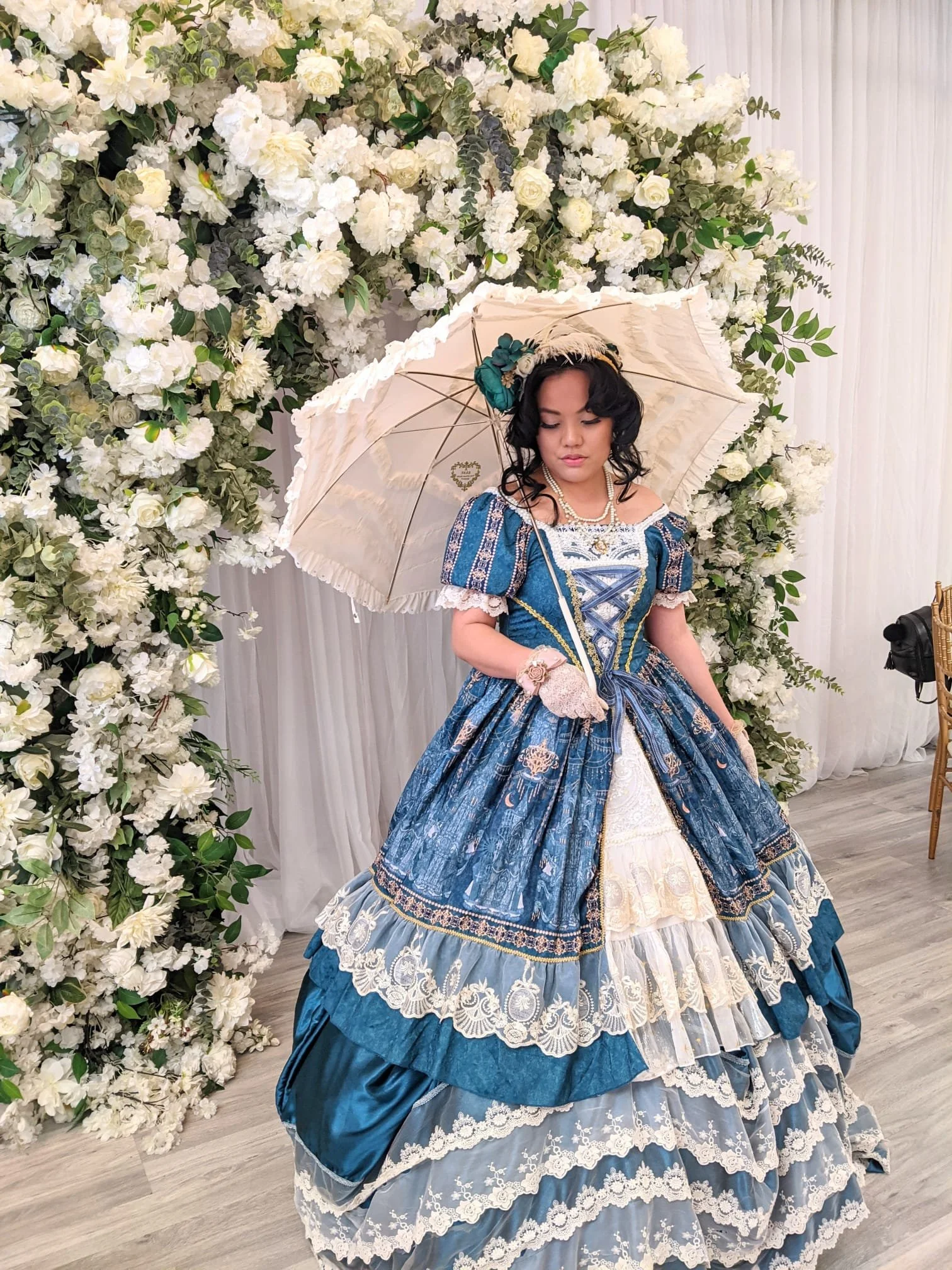 A woman dressed in a vintage, Victorian-style blue and white gown with lace and embroidery details. She holds a lace glove in one hand, wearing a necklace, bracelet, and hat with teal flowers. She stands in front of a large floral backdrop of white r