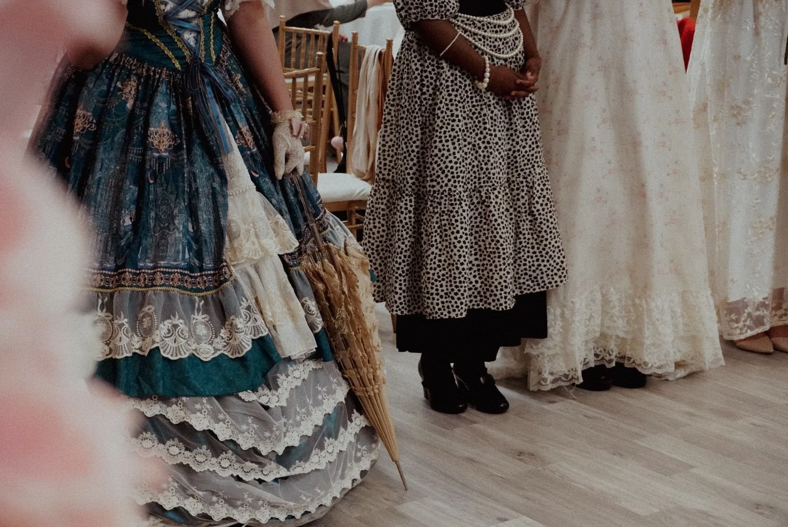 A group of women dressed in vintage and lace dresses, standing on a wooden floor in a room with chairs and a clothing rack in the background.
