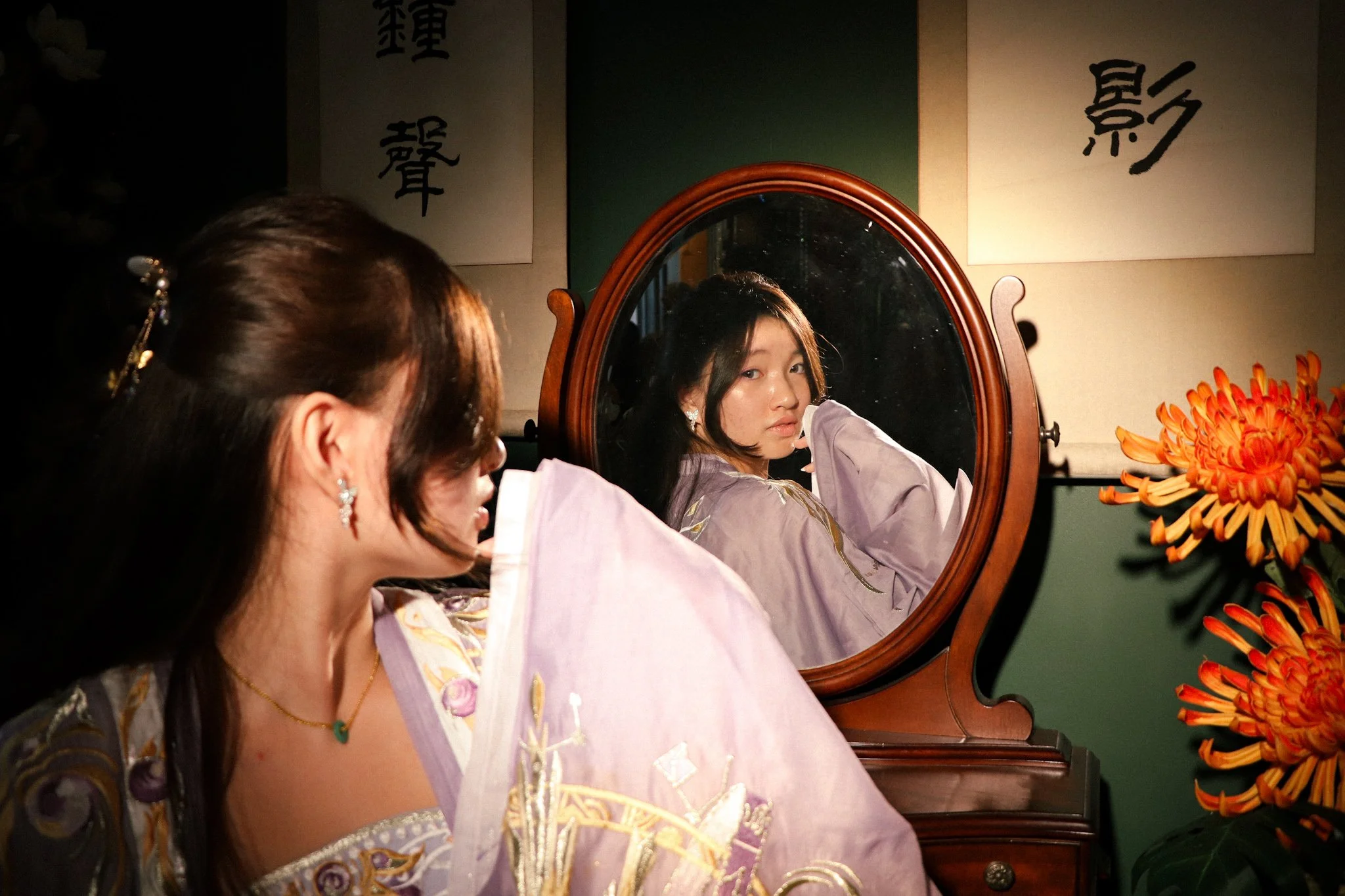 A woman with dark hair looking into a mirror on a vintage wooden dresser. The mirror reflects her face and part of her shoulder, and she is dressed in traditional clothing with floral embroidery. An orange flower arrangement is visible on the right s