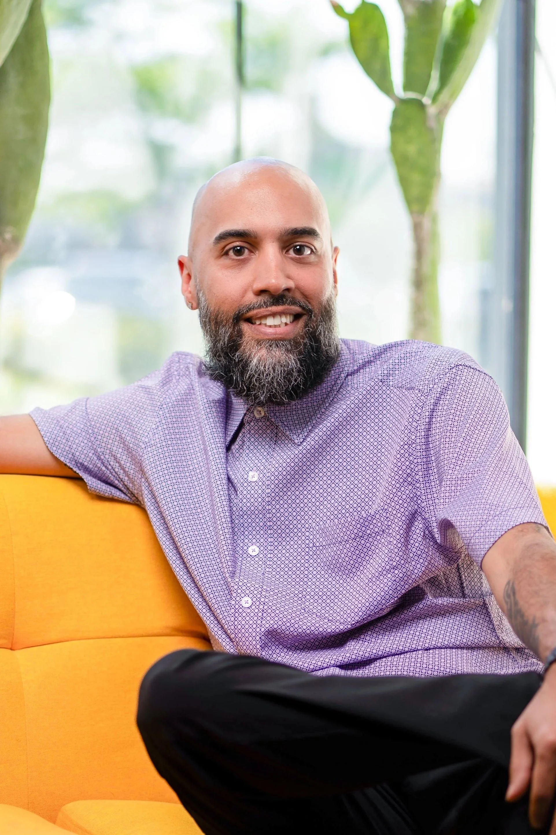 A man in a tuxedo with a bow tie, sporting a neat hairstyle and beard, smiling slightly against a gray background.