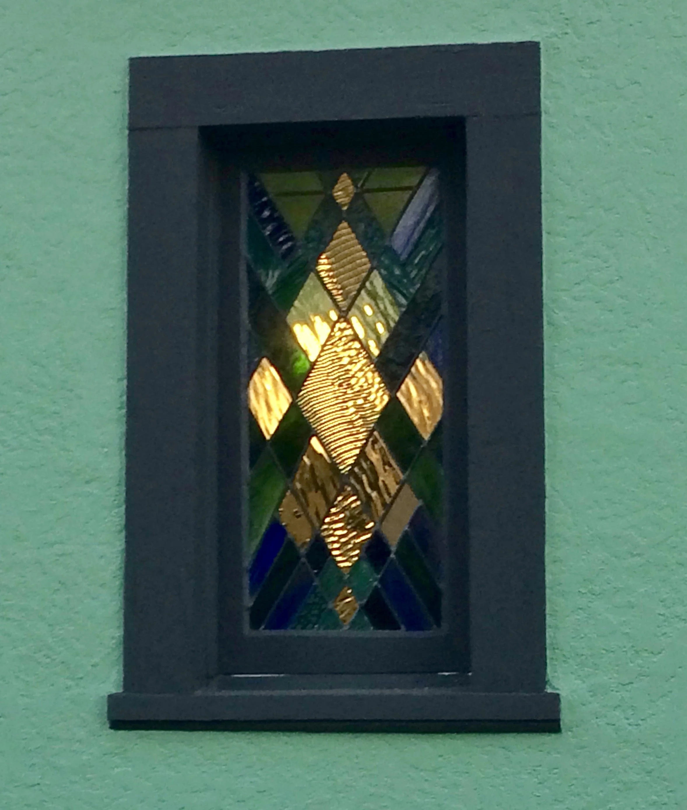 Colorful stained glass window with geometric patterns, framed in black, mounted on a green wall.