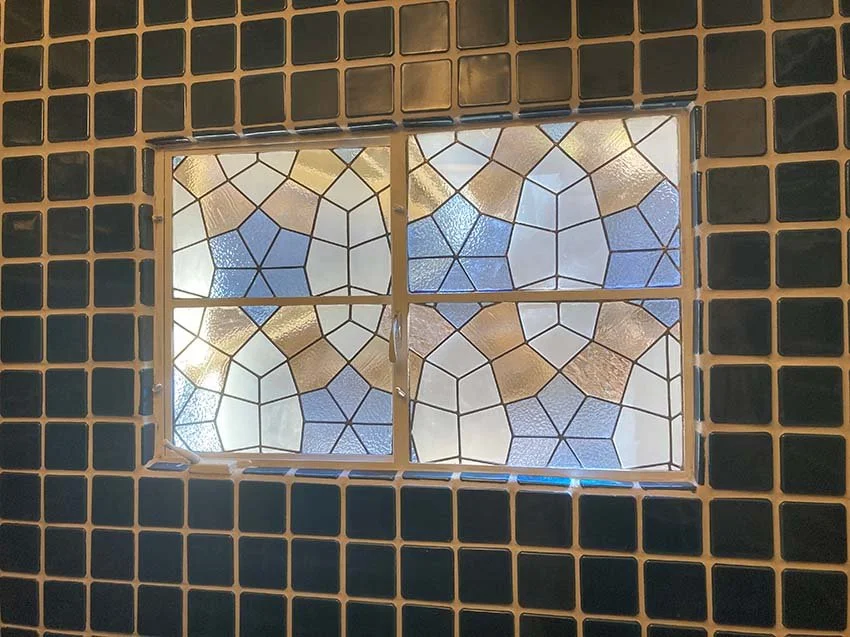 A small window with stained glass panes set in a wall of black tiles with beige grout.
