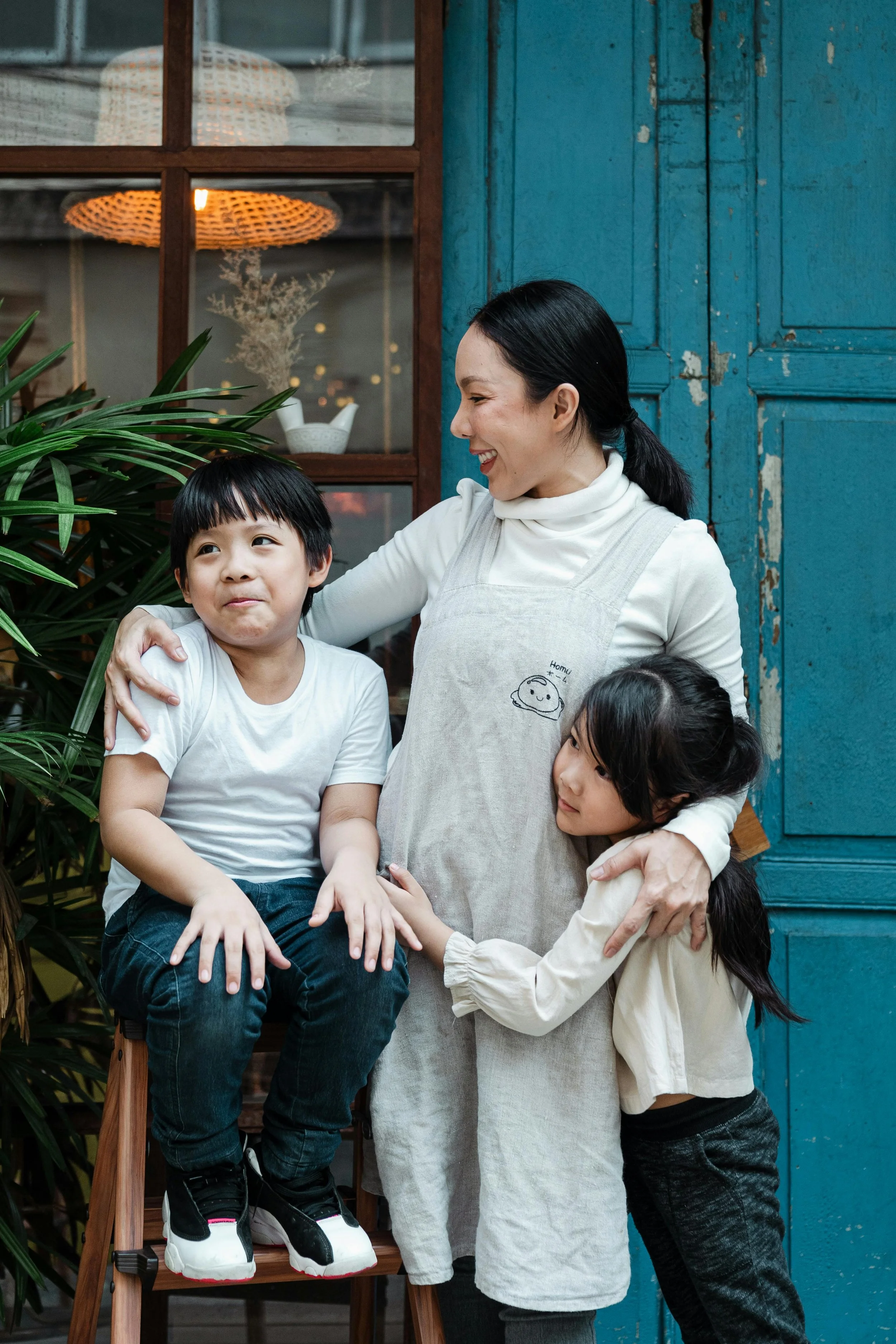 A woman smiling and hugging two children, a boy sitting on a wooden stool and a girl standing, outside near a blue door and green plants.