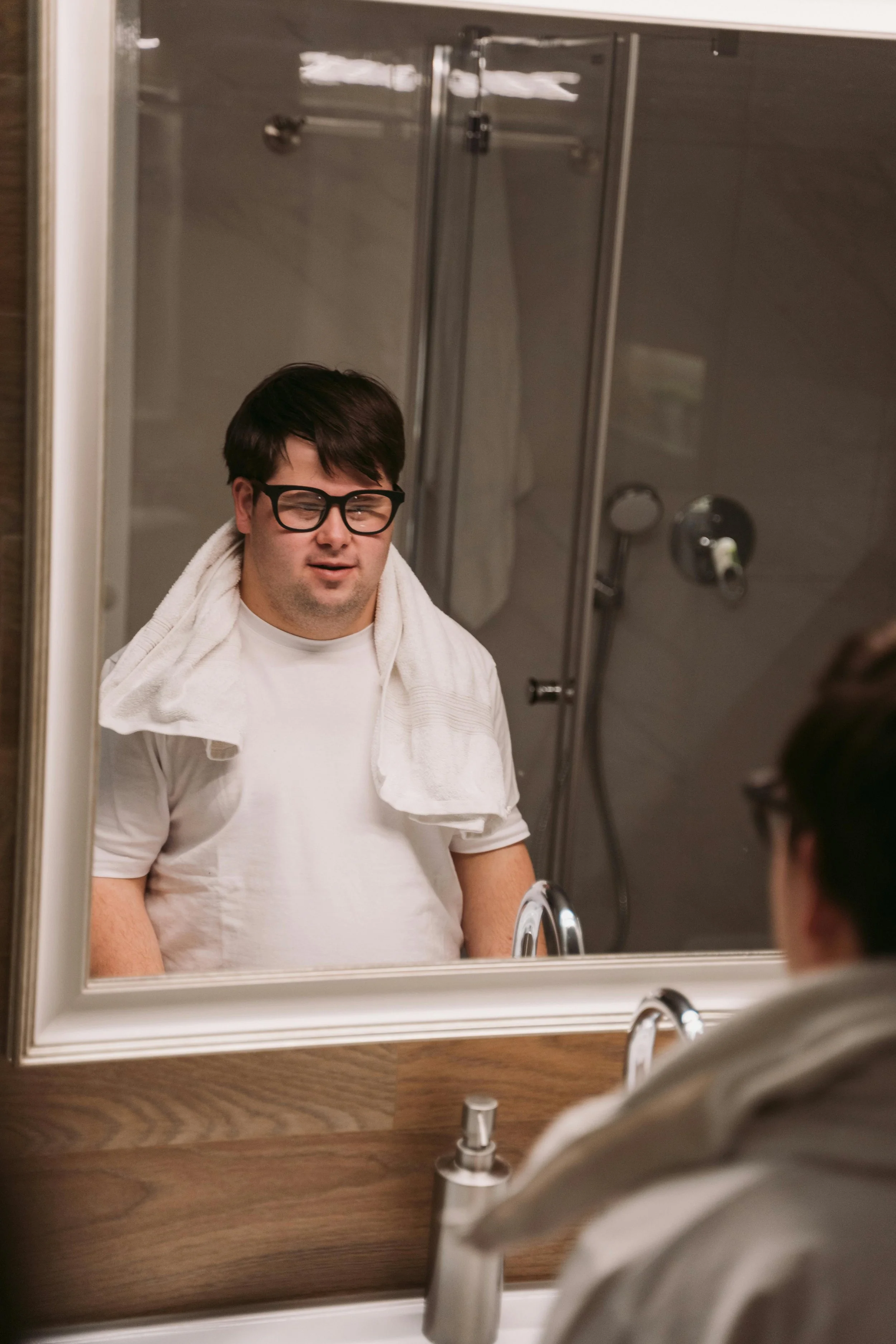 A young man with glasses and dark hair looking at himself in a bathroom mirror, wearing a white t-shirt with a towel draped around his neck, with a shower and bathroom fixtures reflected in the background.