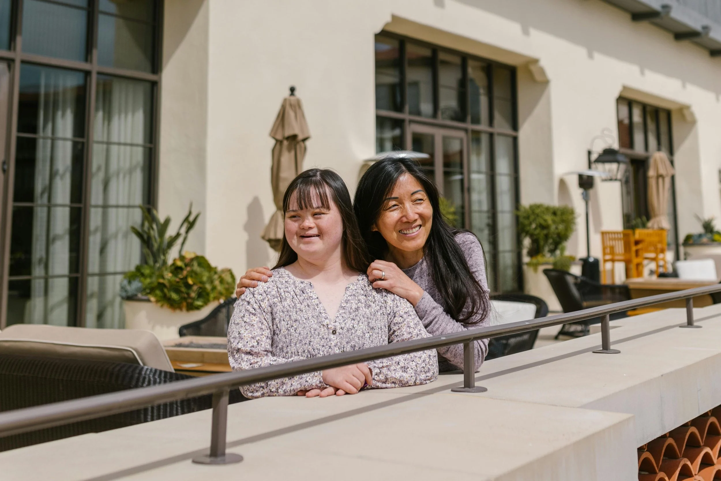 Two women smiling on a patio with seating, large windows, and plants in the background.