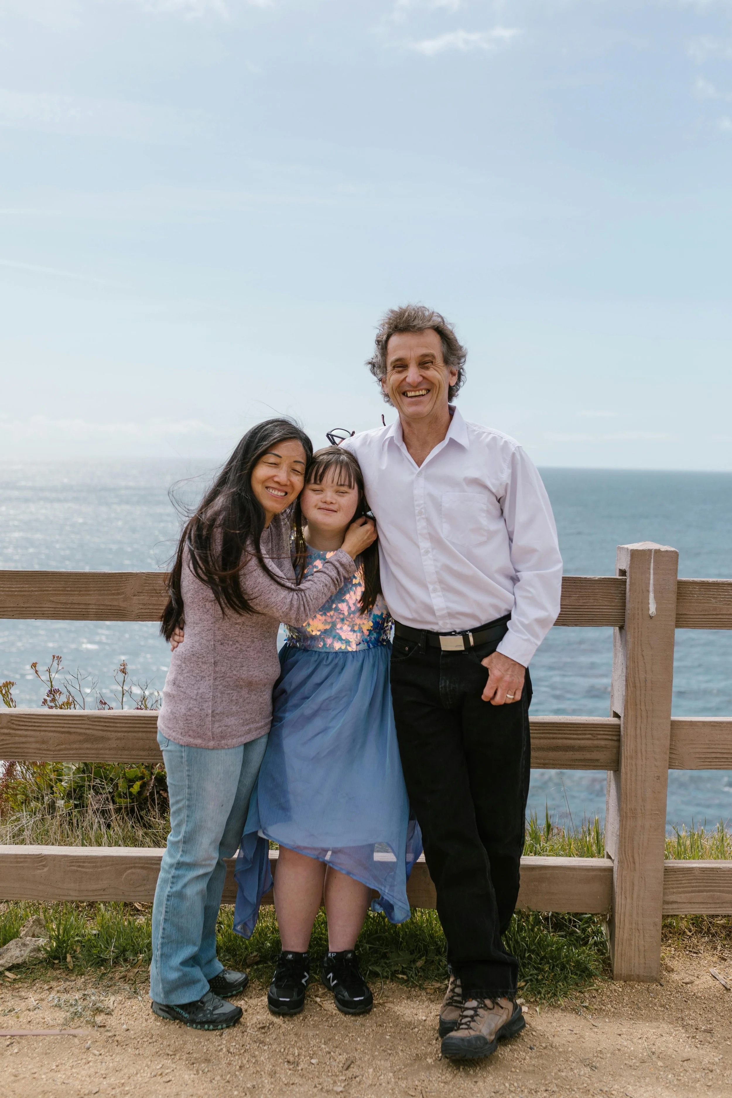 A family of three standing together outdoors near a wooden fence with the ocean and sky in the background. The family consists of a woman, a young girl, and a man, all smiling.