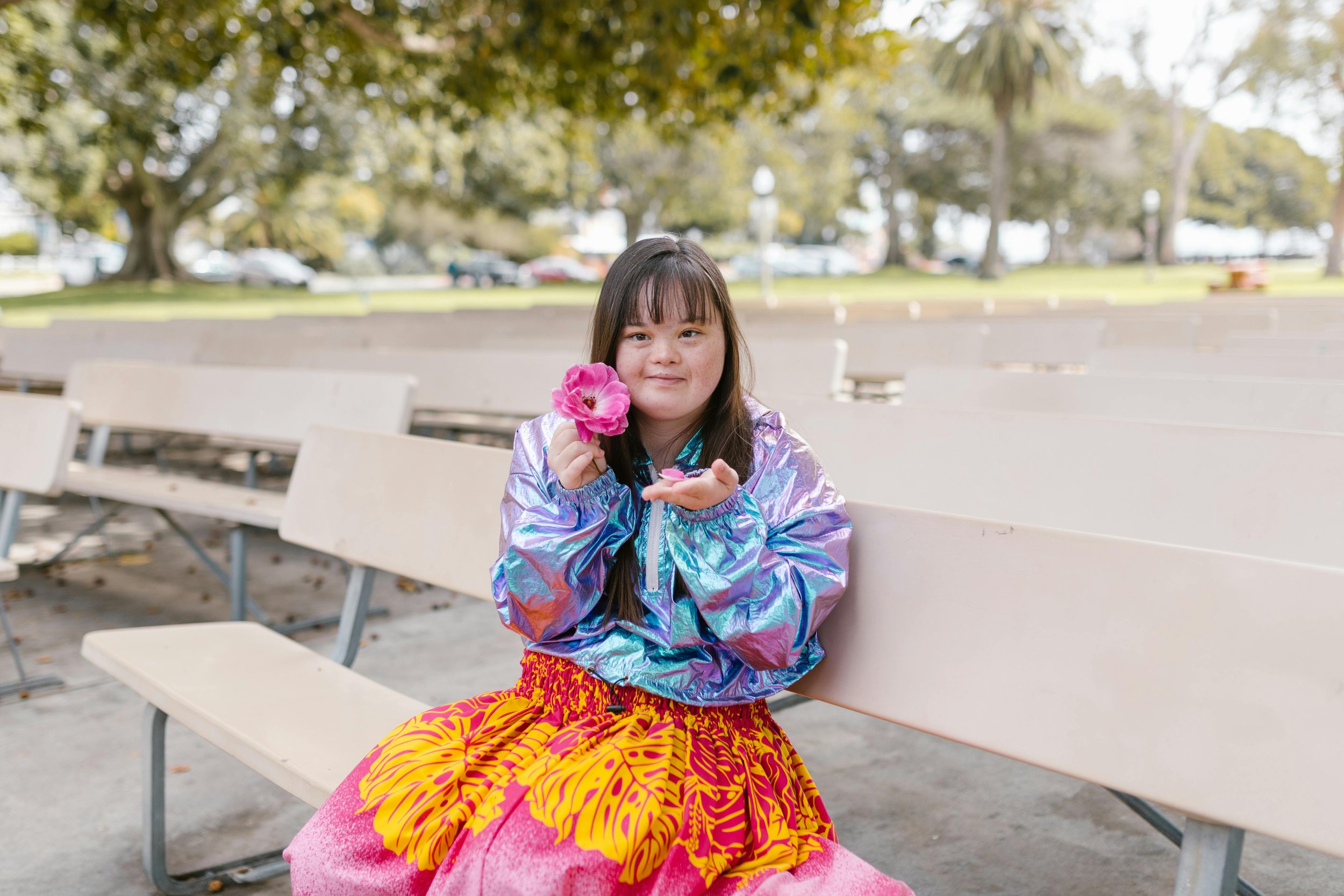 A young girl sitting on a park bench holding a pink flower, wearing a shiny iridescent jacket and a vibrant yellow and pink skirt, with trees and park area in the background.