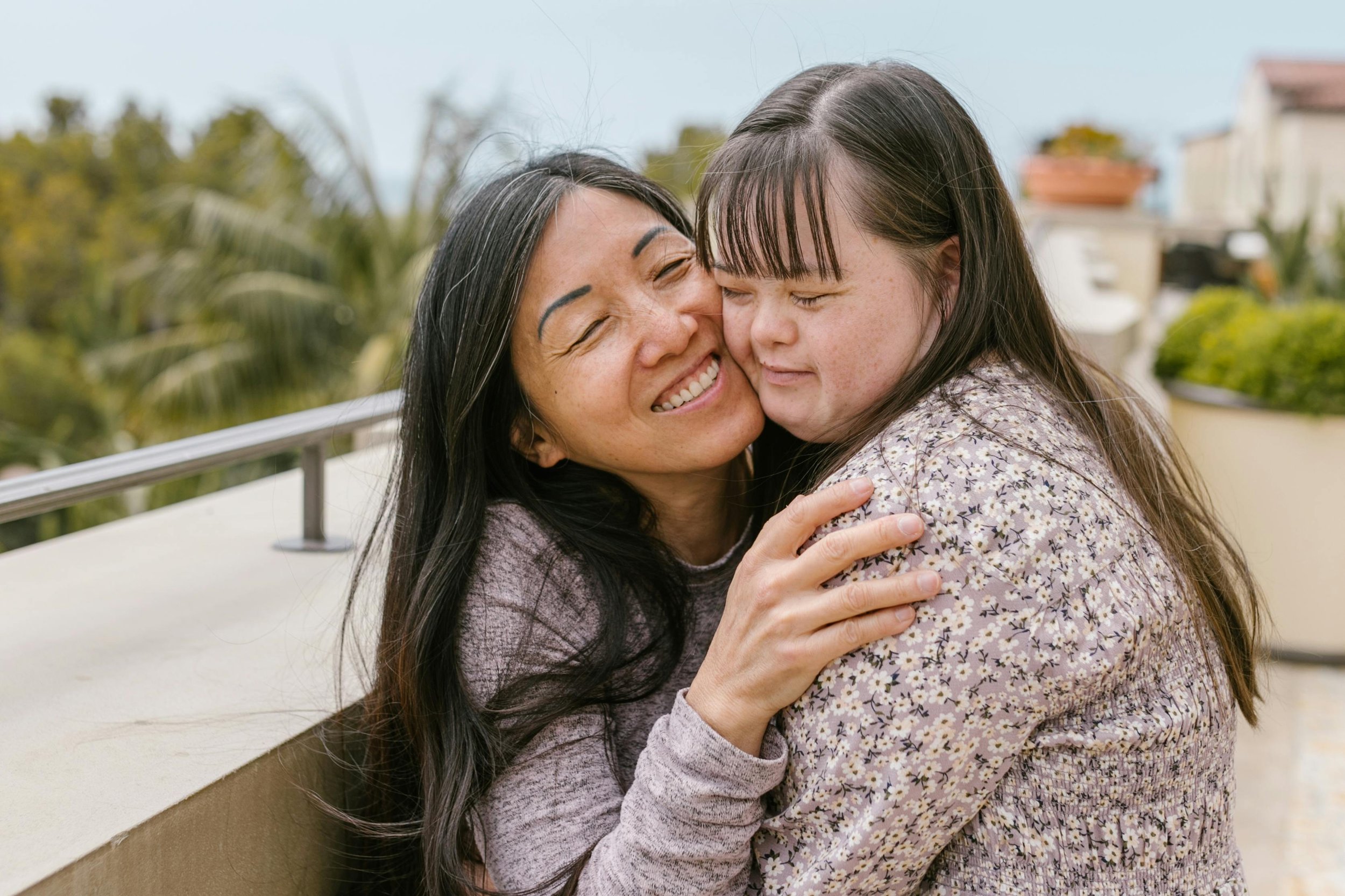 A woman with long dark hair and a floral blouse hugs a young girl with straight dark hair and bangs, both smiling warmly with eyes closed outside on a balcony or terrace.