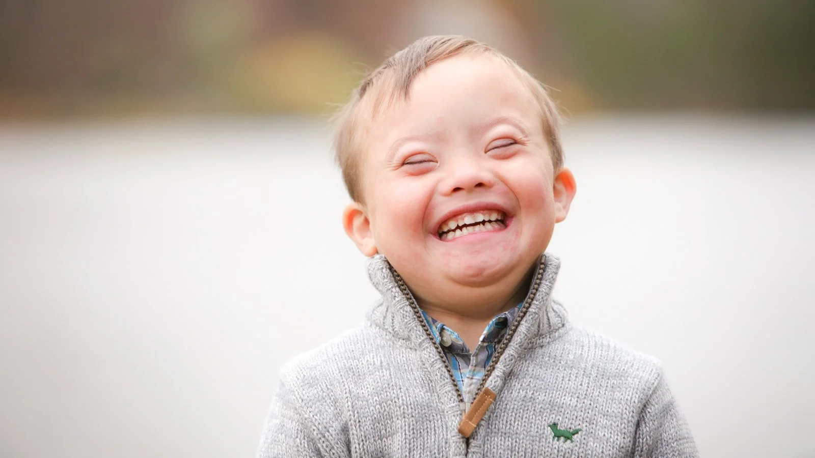 A young boy smiling widely with his eyes closed, wearing a gray sweater and a plaid shirt, standing outdoors with a blurred natural background.