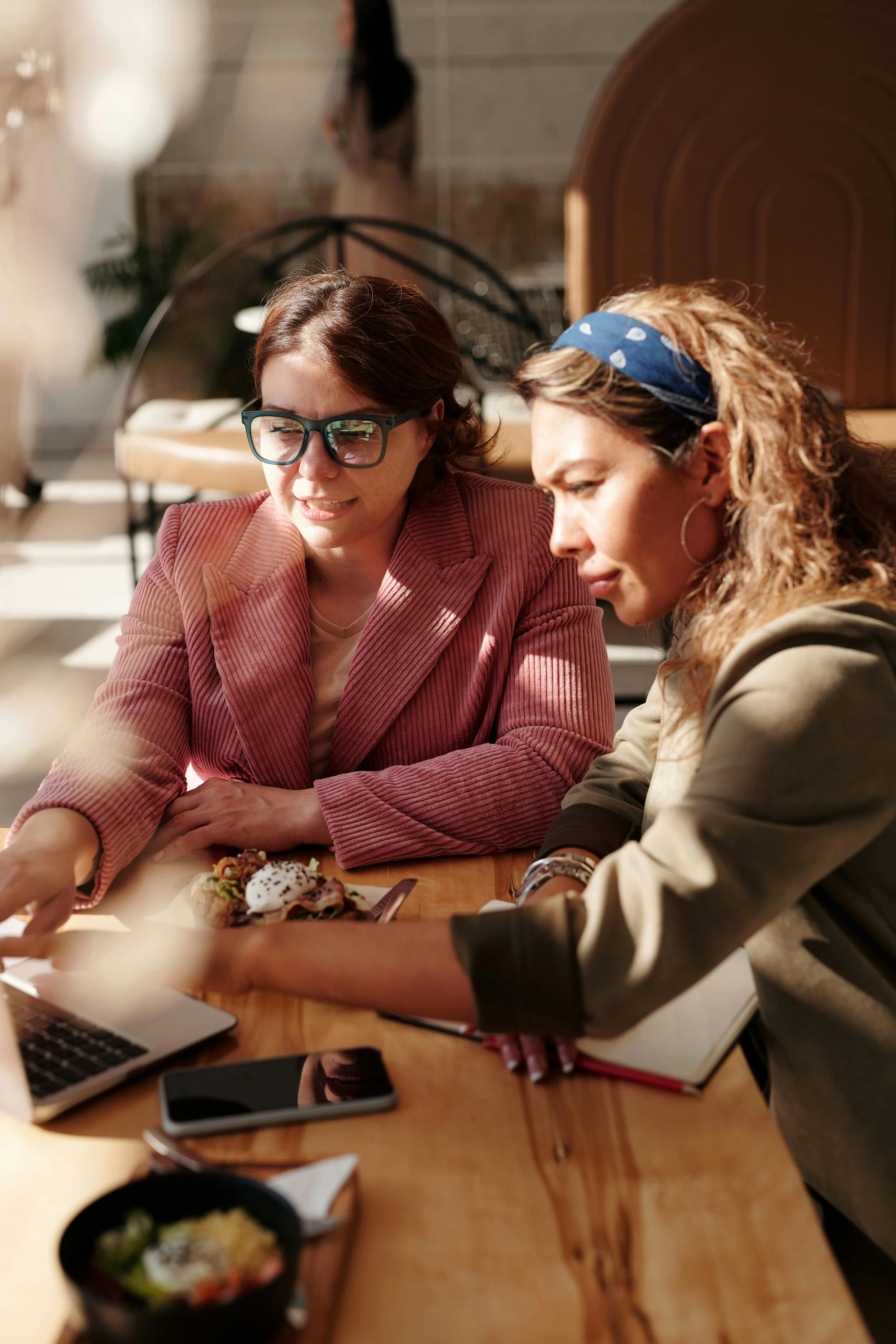 Two women sitting at a wooden table in a bright cafe, looking at a laptop and a phone, with a small bowl of salad, a plate with dessert, and a notebook on the table.