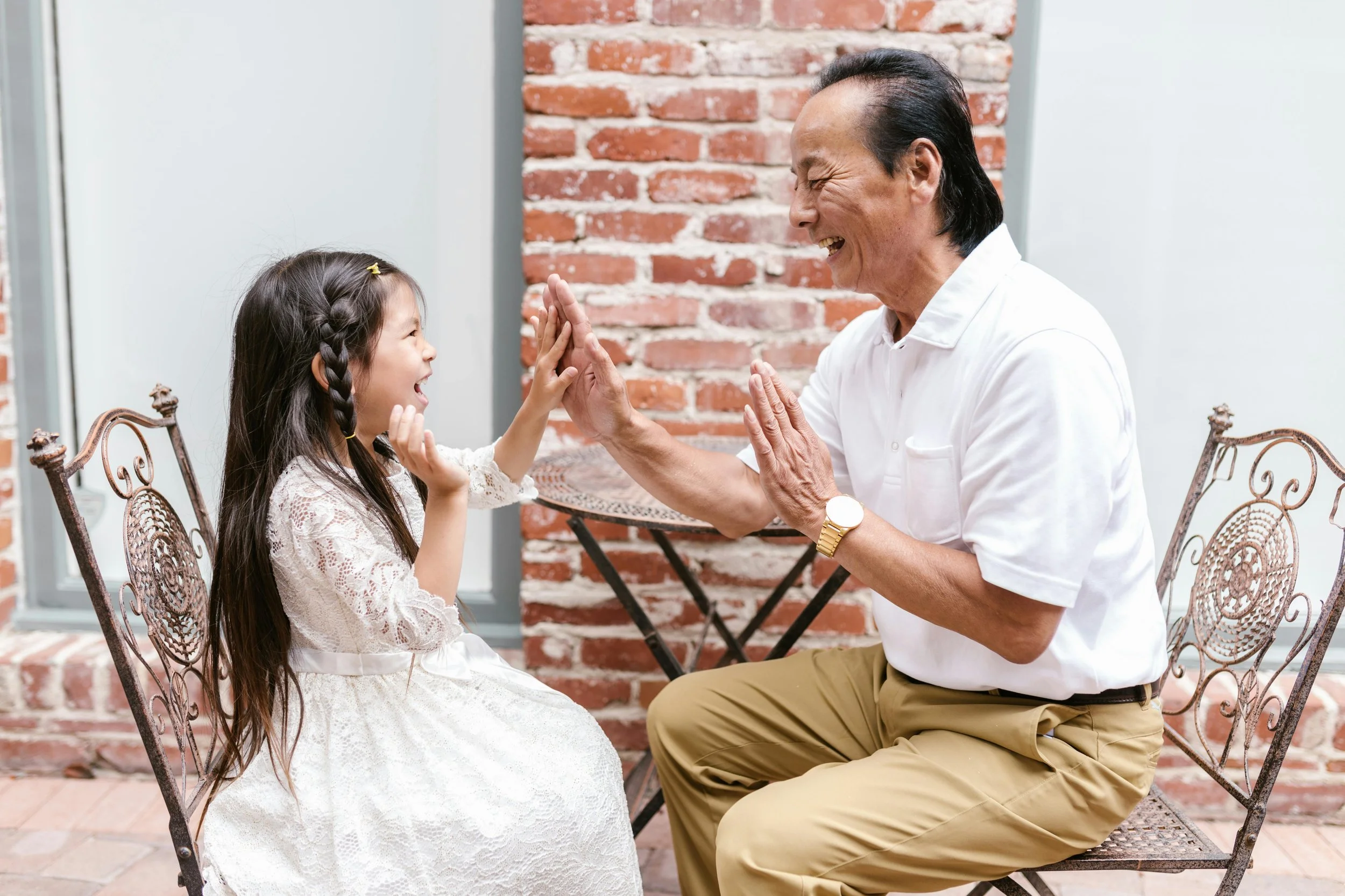 A young girl and an elderly man are sitting outside, smiling and touching hands in a playful, joyful moment.