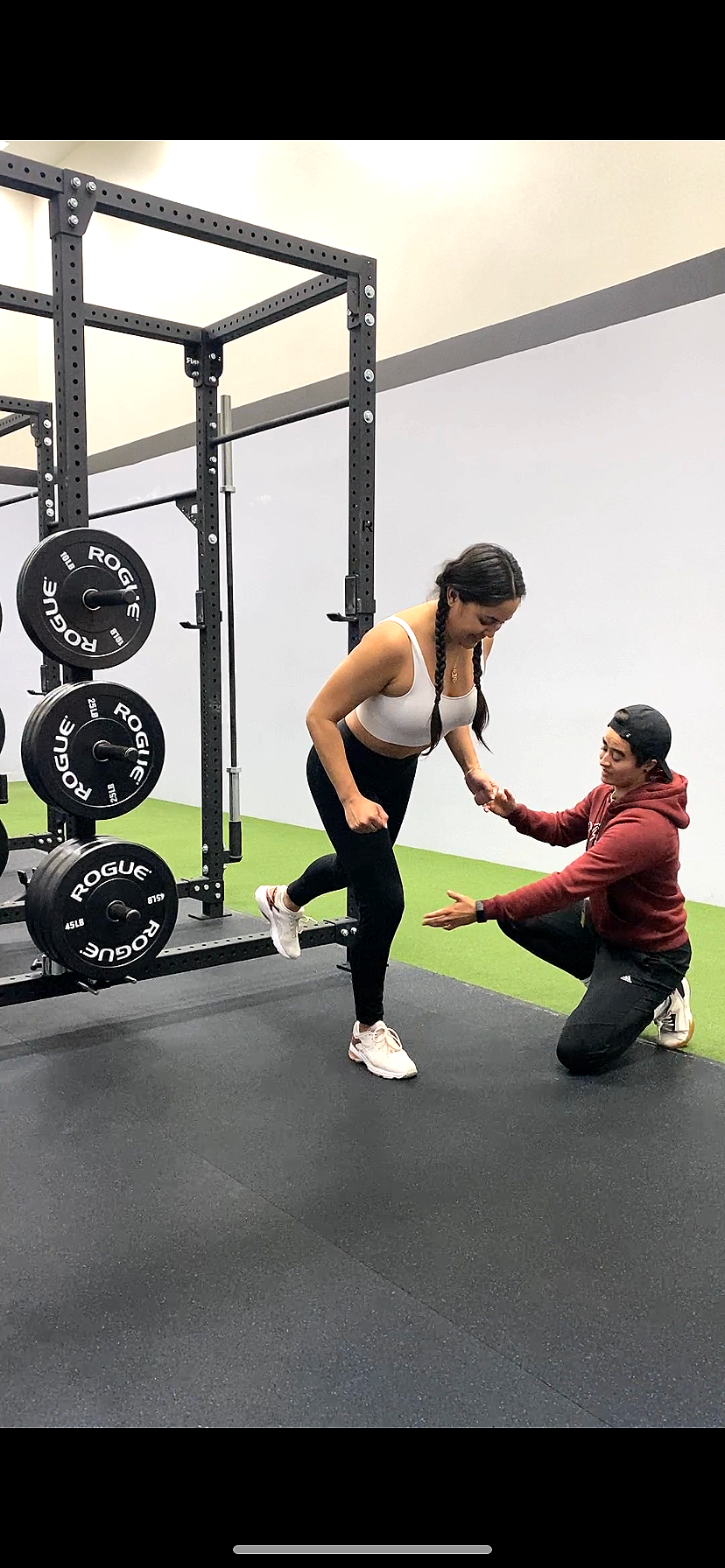 Personal trainer coaching a client through a single-leg squat in a gym, providing hands-on support to ensure proper form and balance. Strength training session focused on stability and functional movement.