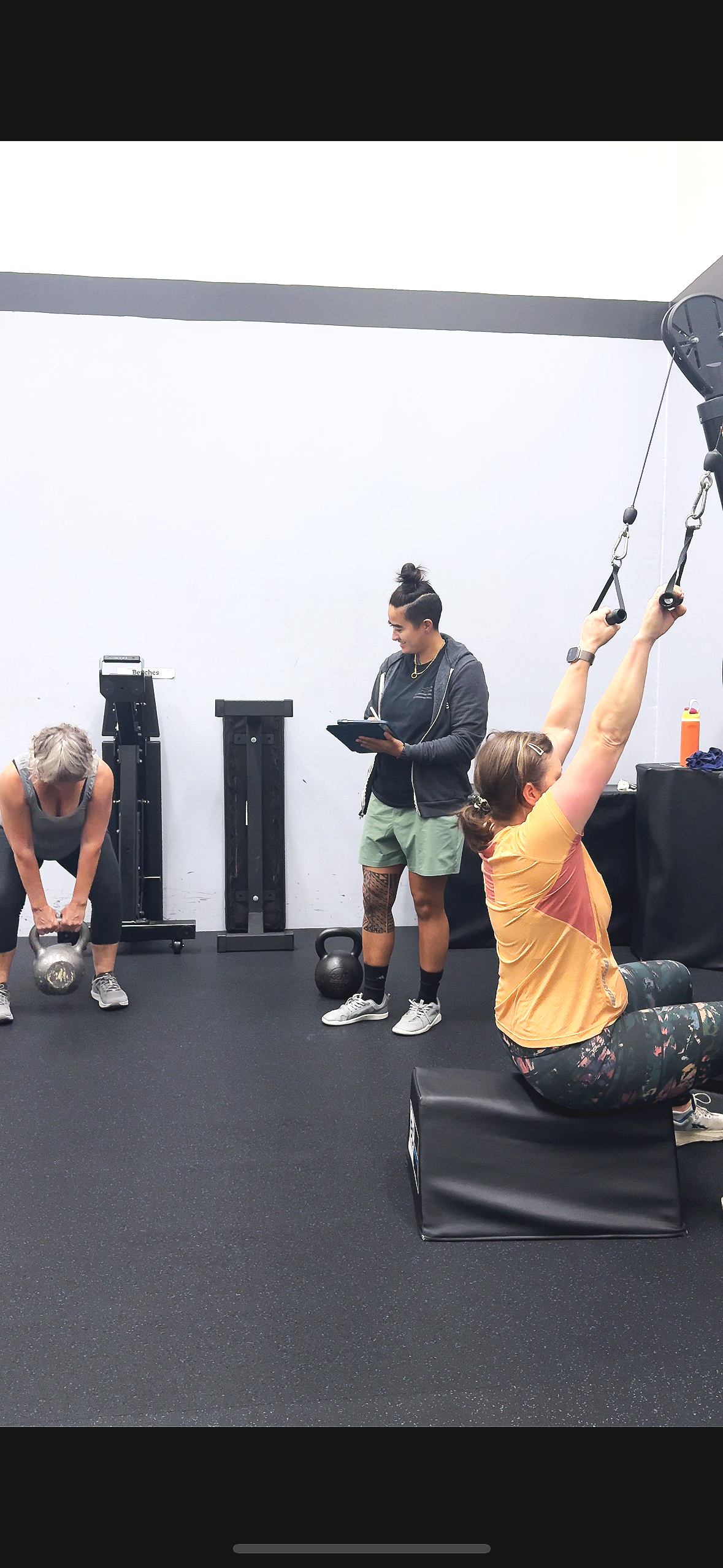 Personal trainer coaching a client on a cable machine exercise, providing hands-on guidance to ensure proper form and alignment. The client is in a strong stance, focusing on controlled movement. A client in the background performs kettlebell exercis