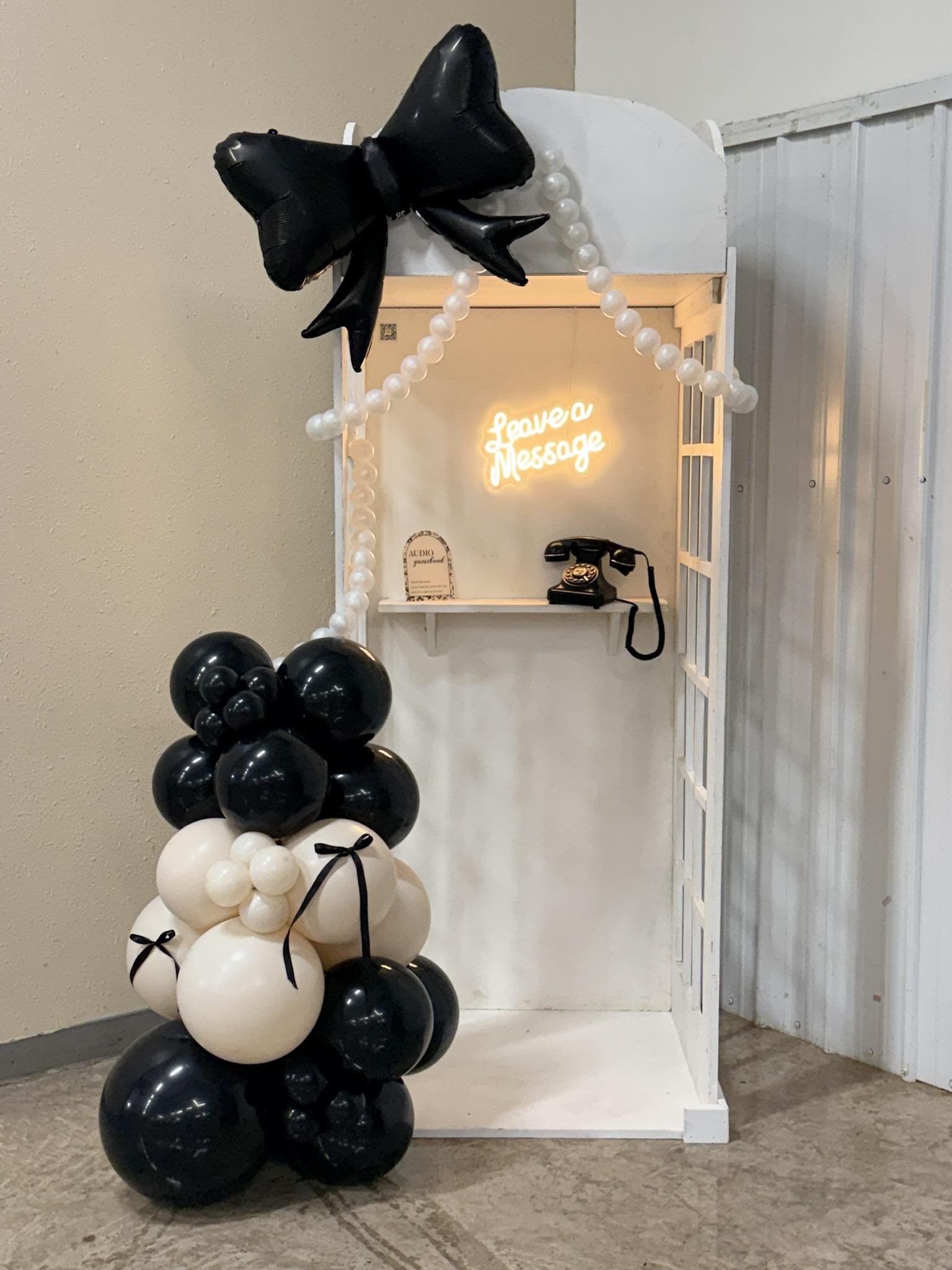 A decorative telephone booth with a neon sign that says 'Leave a Message' inside, an old-fashioned rotary phone, and a black-and-white patterned floor.