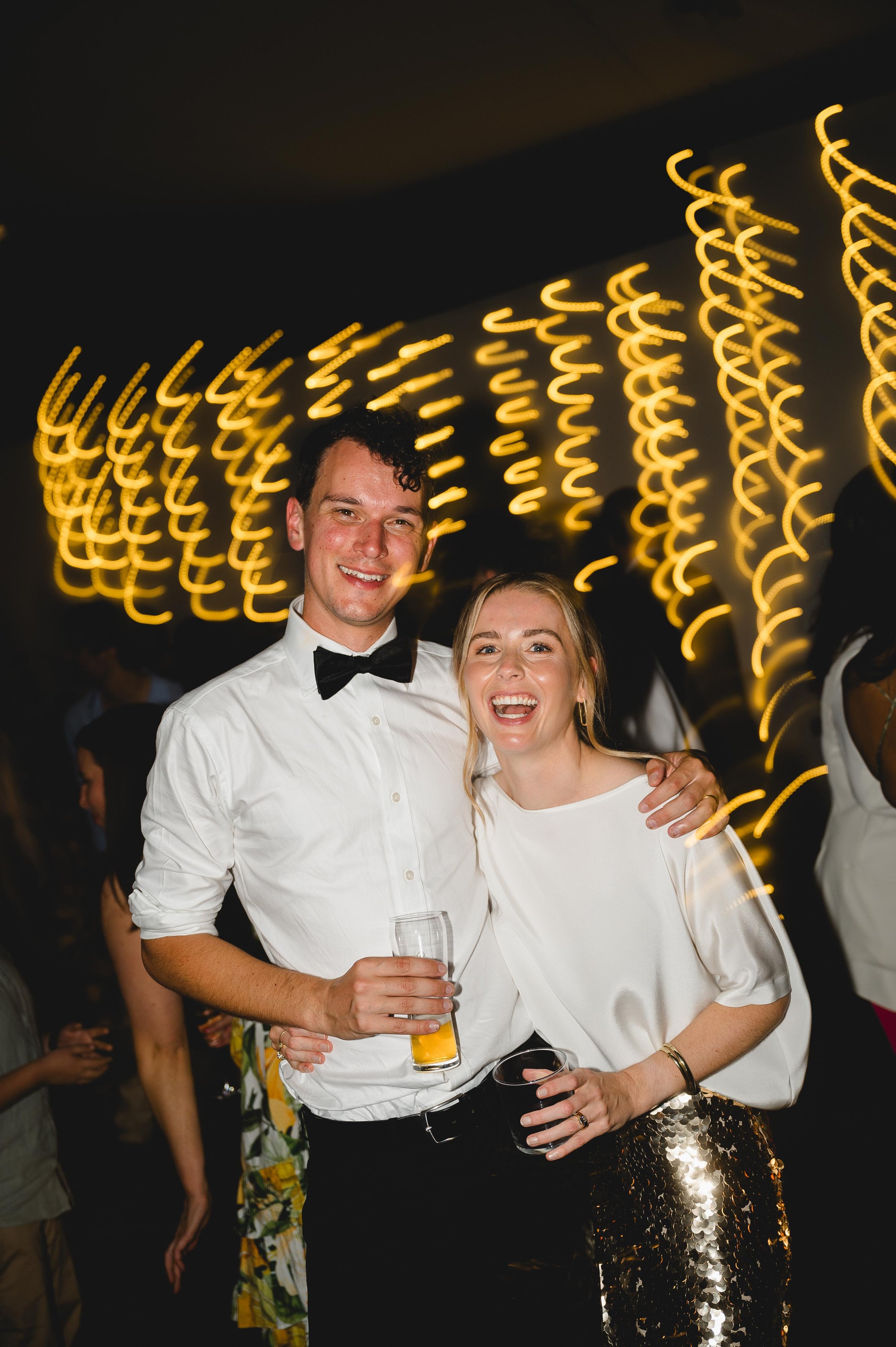 A smiling couple poses with drinks at a party with decorative lights in the background.