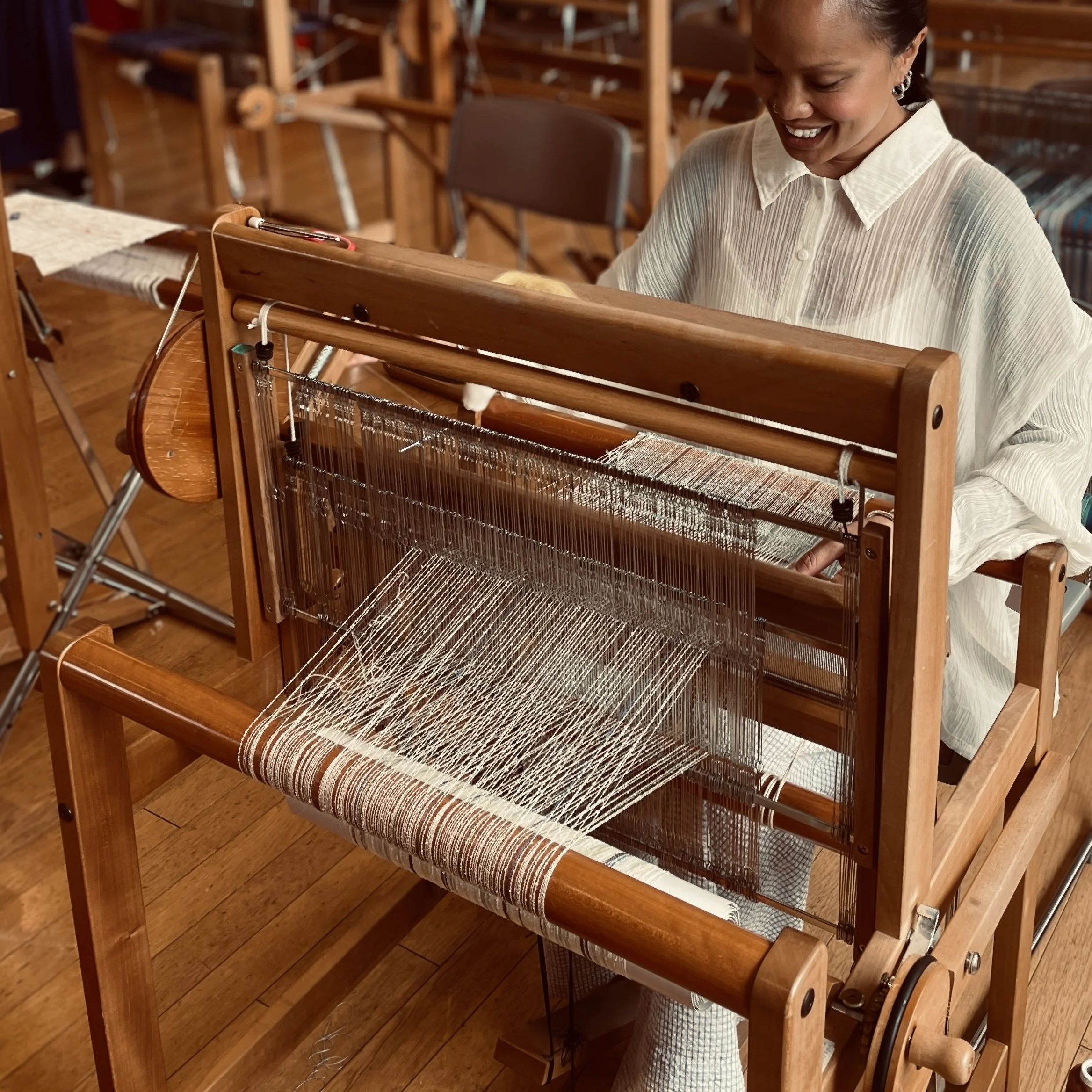 A woman smiling while working on a wooden loom in a room with wooden flooring and furniture.