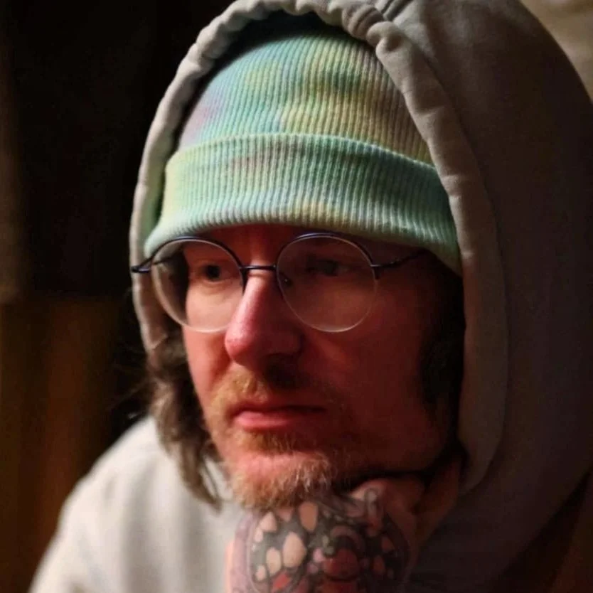 A man with glasses, a rainbow-colored beanie, and a gray hoodie, resting his chin on his hand and looking to the side.